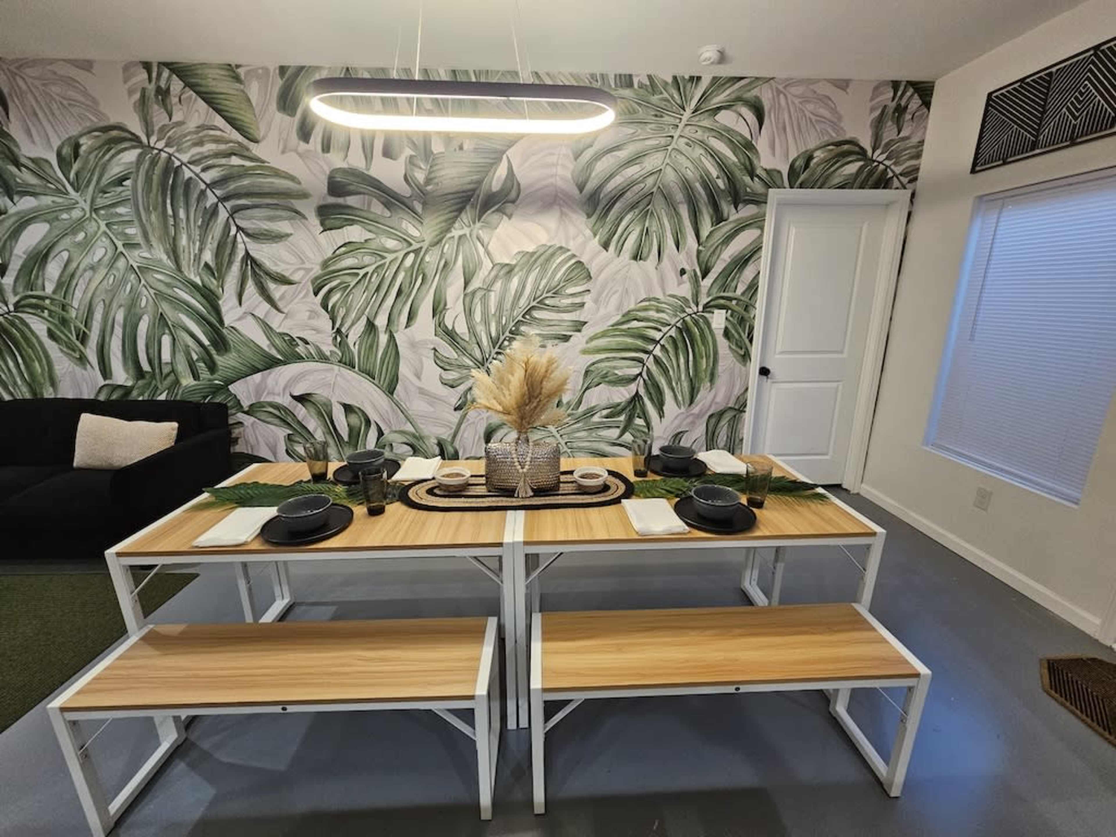 A dining area features a table with a natural wood surface, black plates, and decorative elements, set against a tropical leaf-patterned wallpaper.