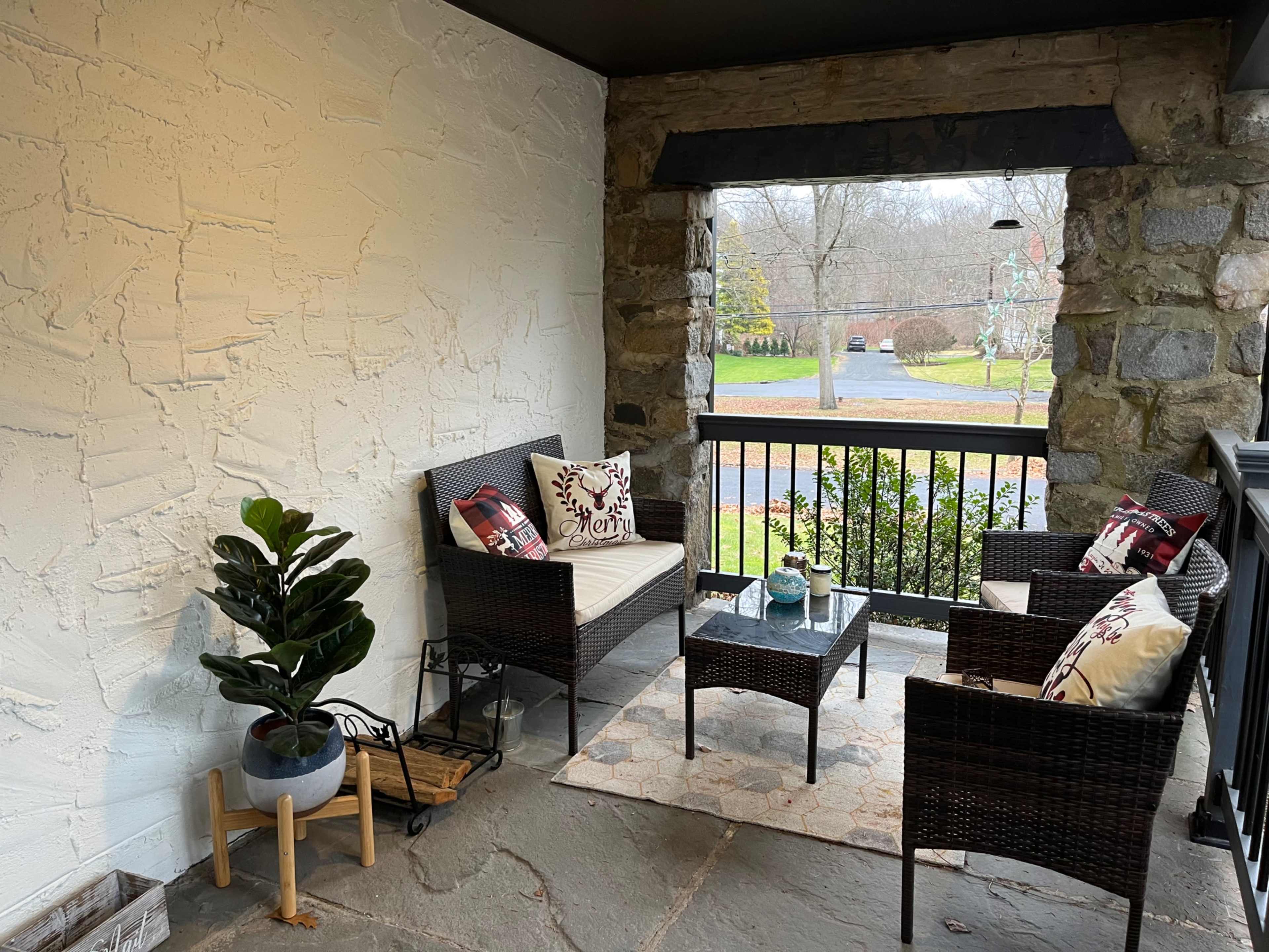 The image shows a cozy porch with wicker furniture, a small coffee table, and a potted plant, set against a stone wall and an outdoor view of a green lawn.