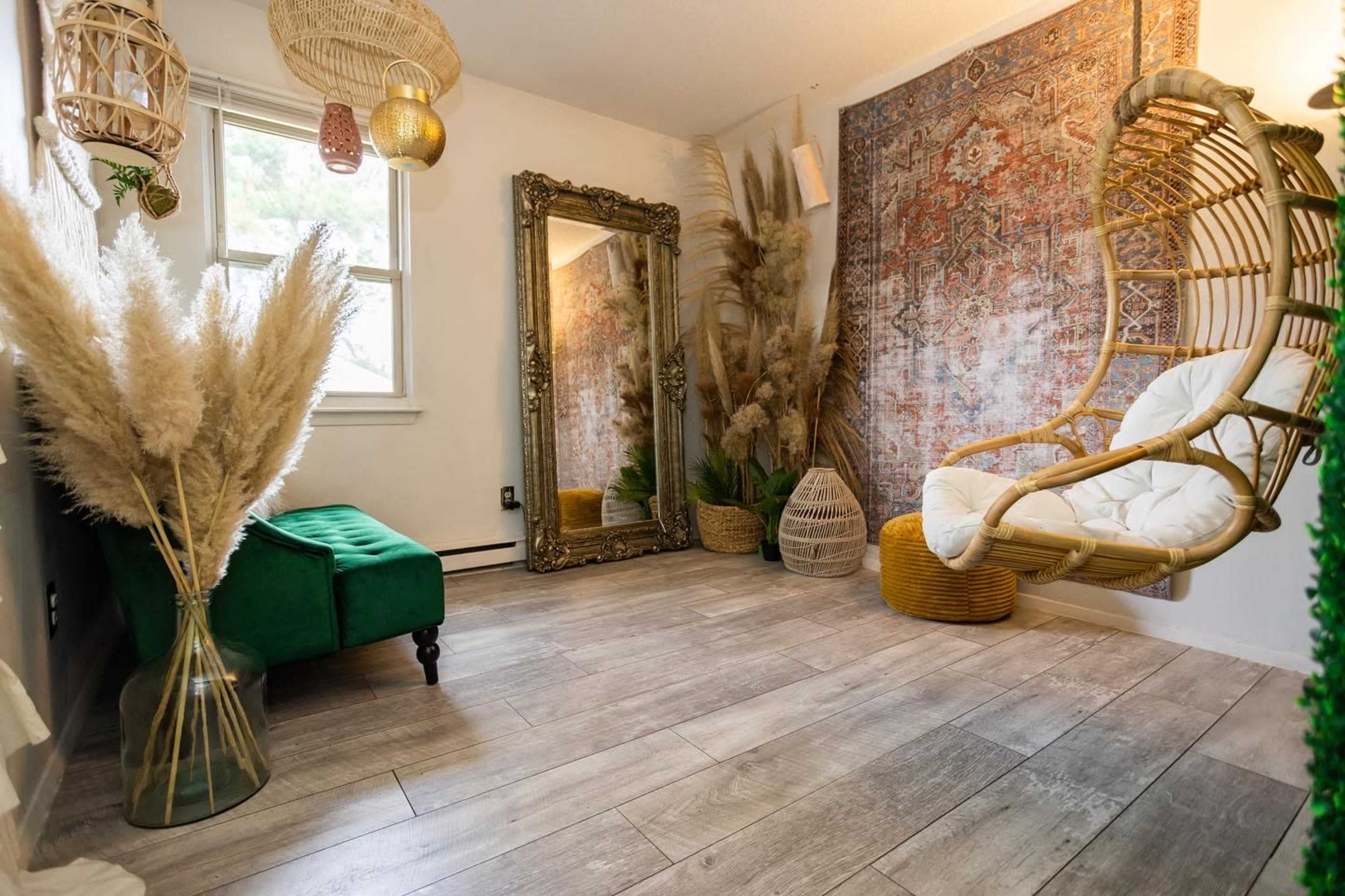 A bright room featuring a hanging chair, a decorative mirror, potted plants, and a green velvet chair, with a patterned wall tapestry and wooden flooring.