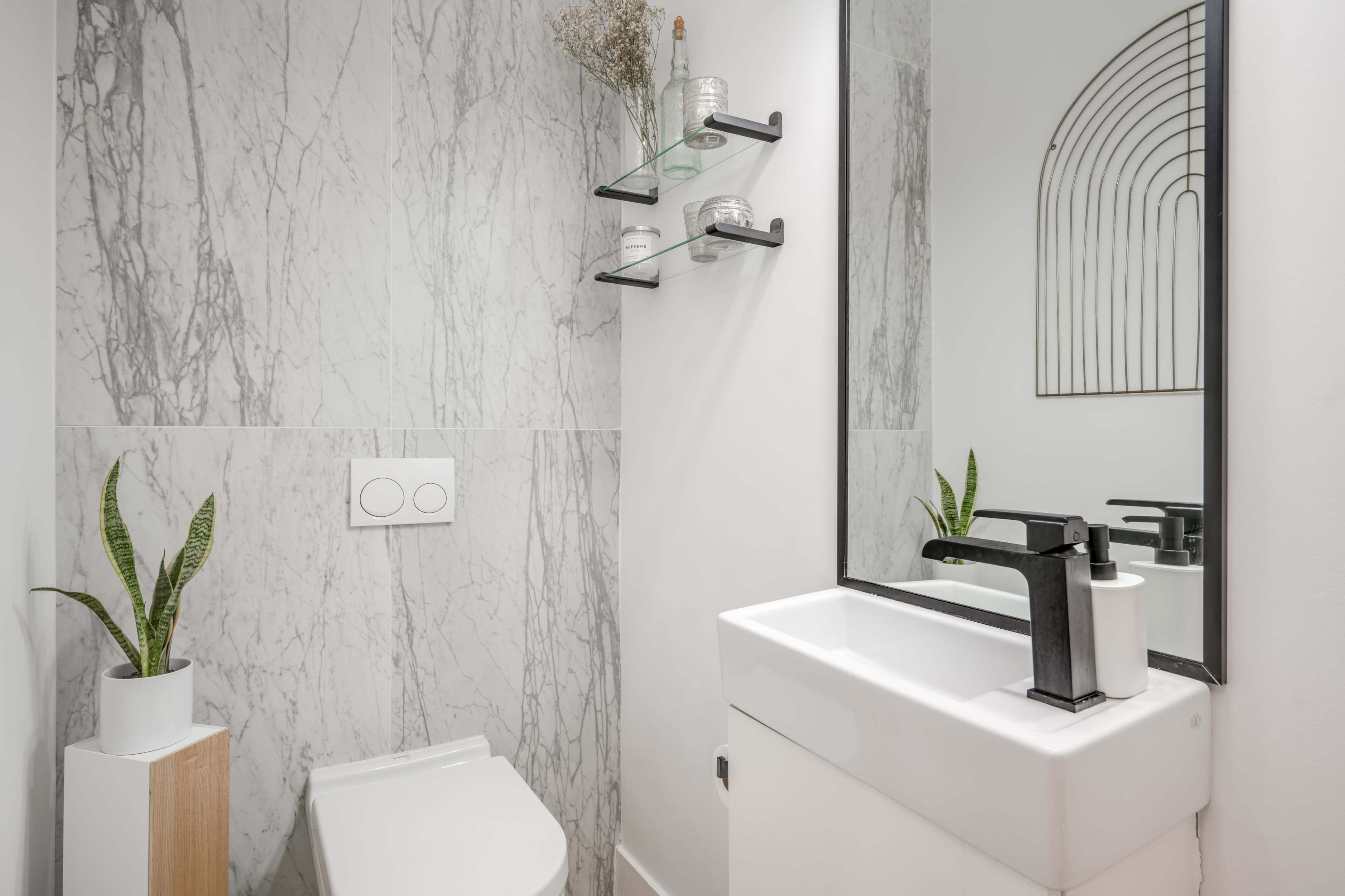 A modern bathroom features a white sink, a black faucet, a wall-mounted mirror, gray marble walls, and a plant in a pot.