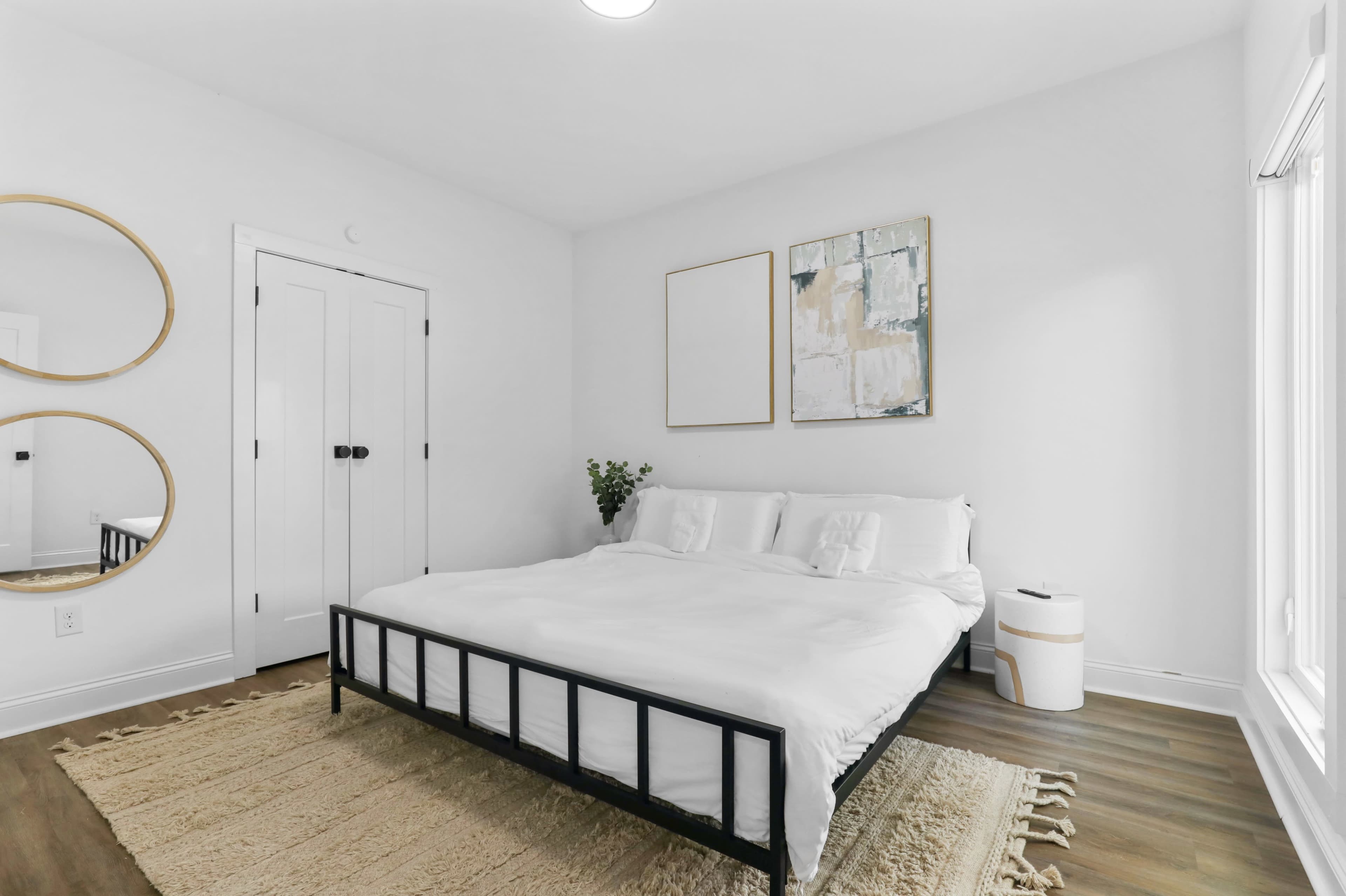 A simple bedroom features a black metal bed frame with a white duvet, framed artwork on the walls, and a mirror beside a closed door.