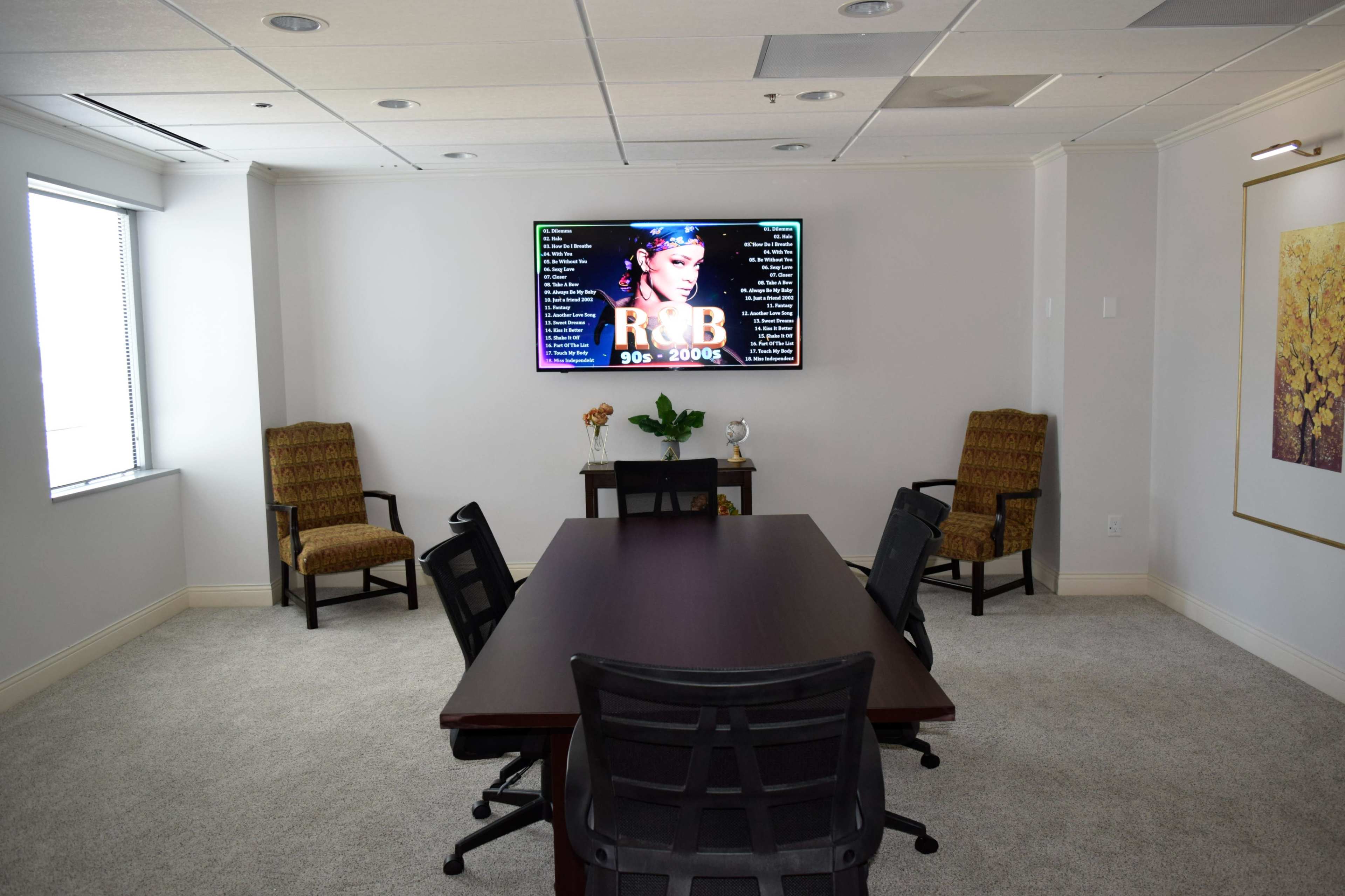 A conference room features a long wooden table surrounded by black office chairs, with a TV displaying R&B music from the 90s and 2000s on the wall.