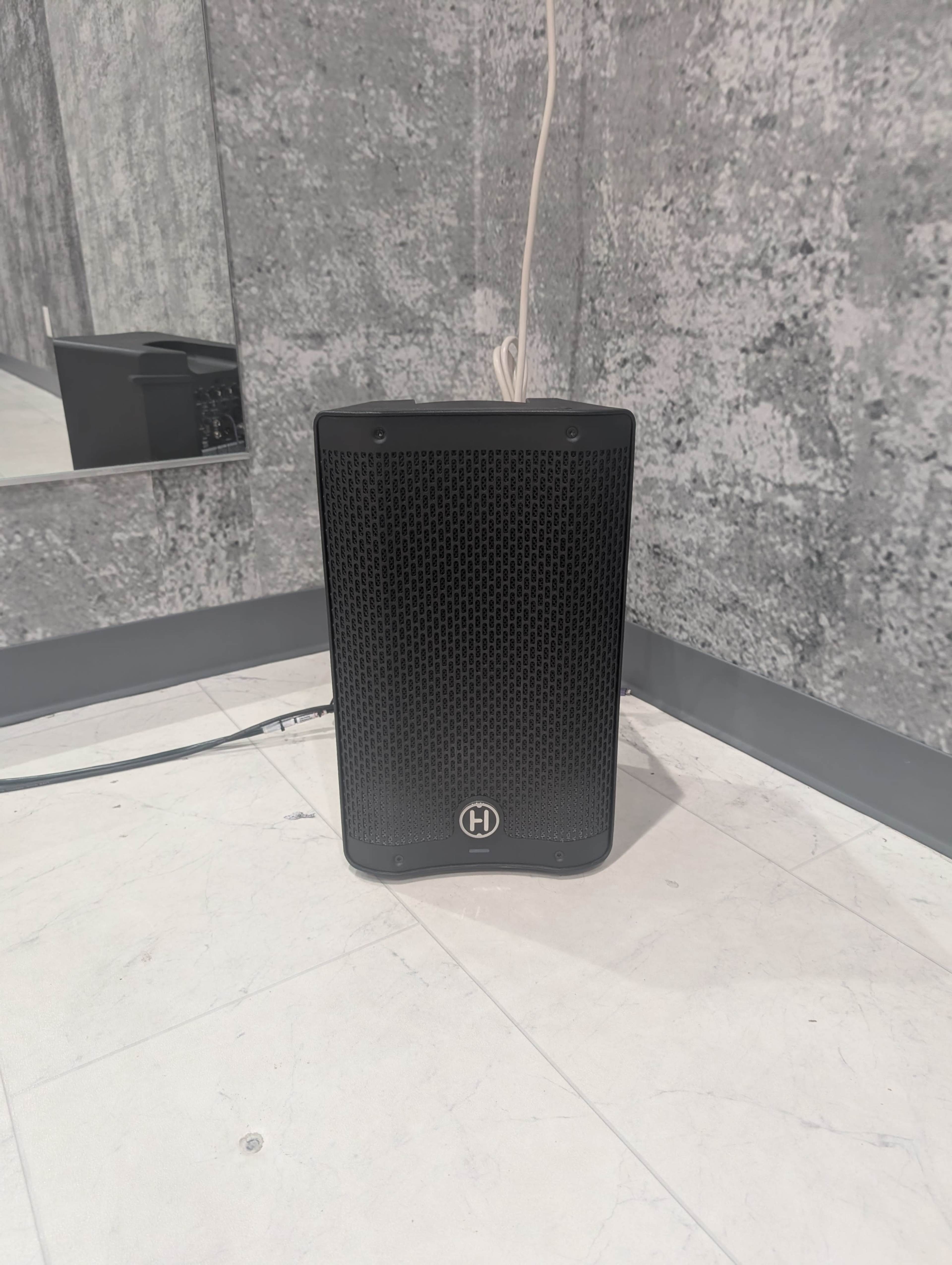A black speaker is positioned in the corner of a room with gray walls and a light-colored floor.