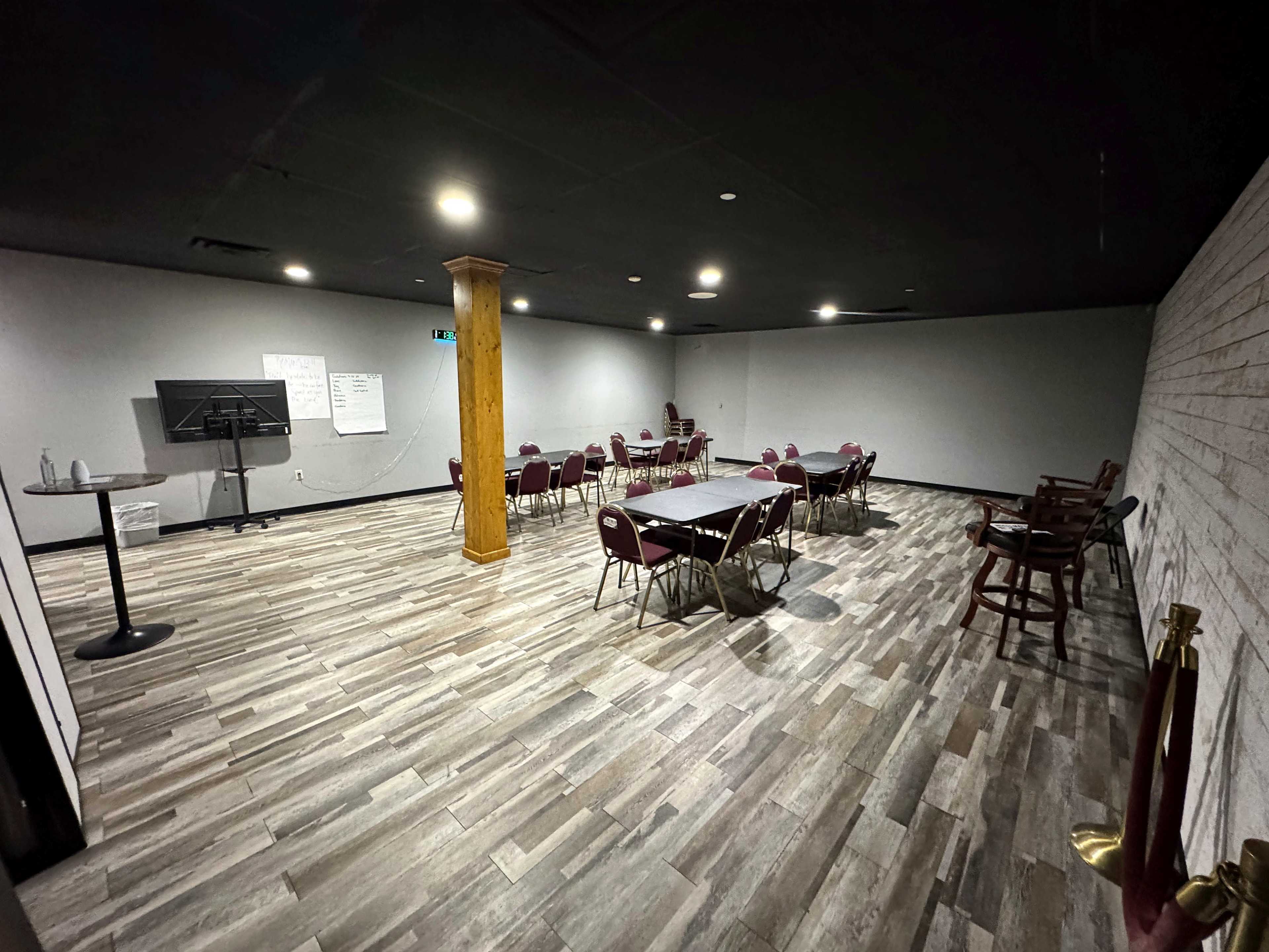 The image shows a meeting room with several tables and chairs arranged in a spacious, well-lit area featuring wooden flooring and a large flat-screen display.