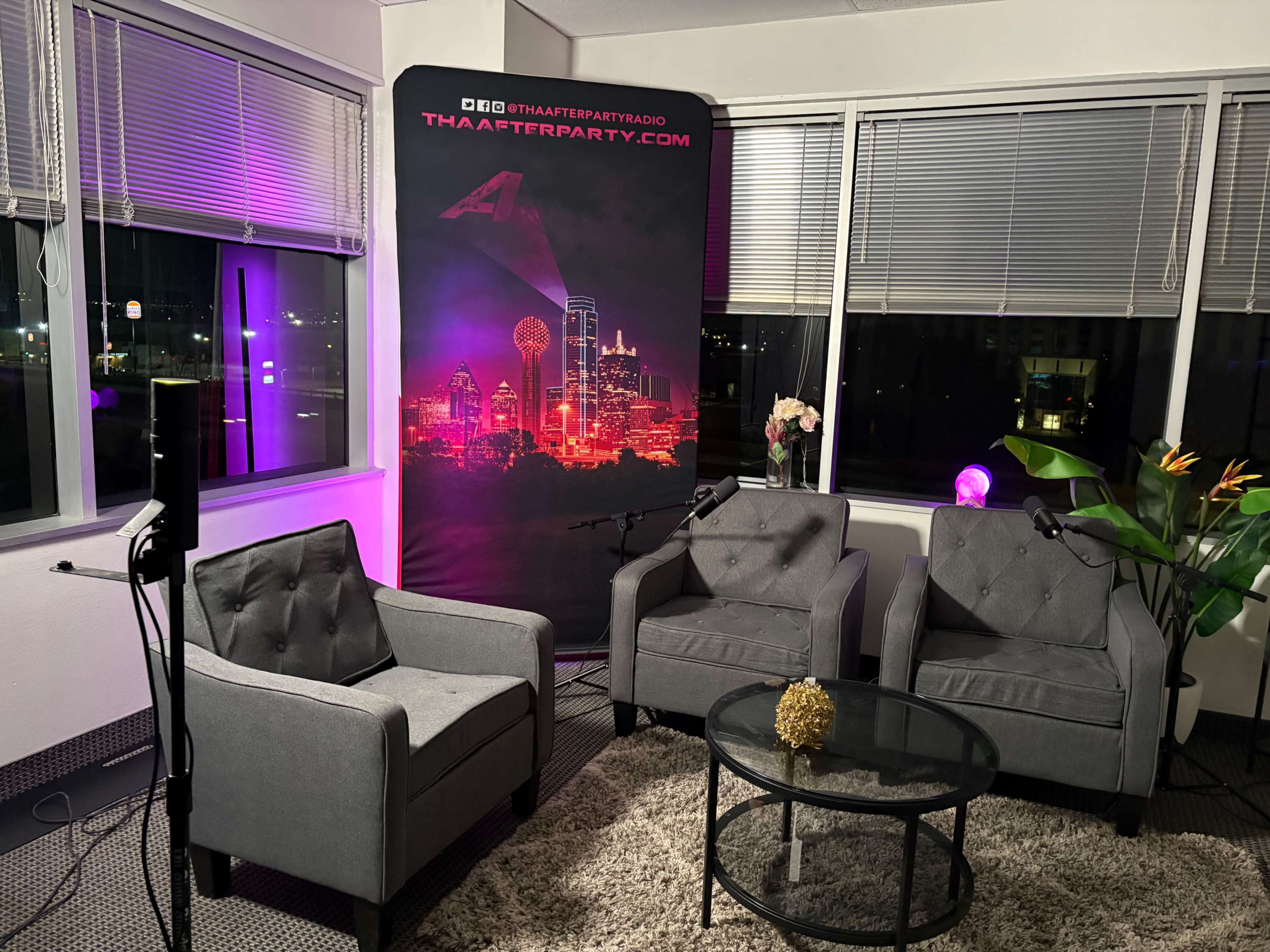 The image shows a cozy studio setup with three gray chairs arranged around a glass table, illuminated by colorful lighting and featuring a backdrop with a skyline and a logo.