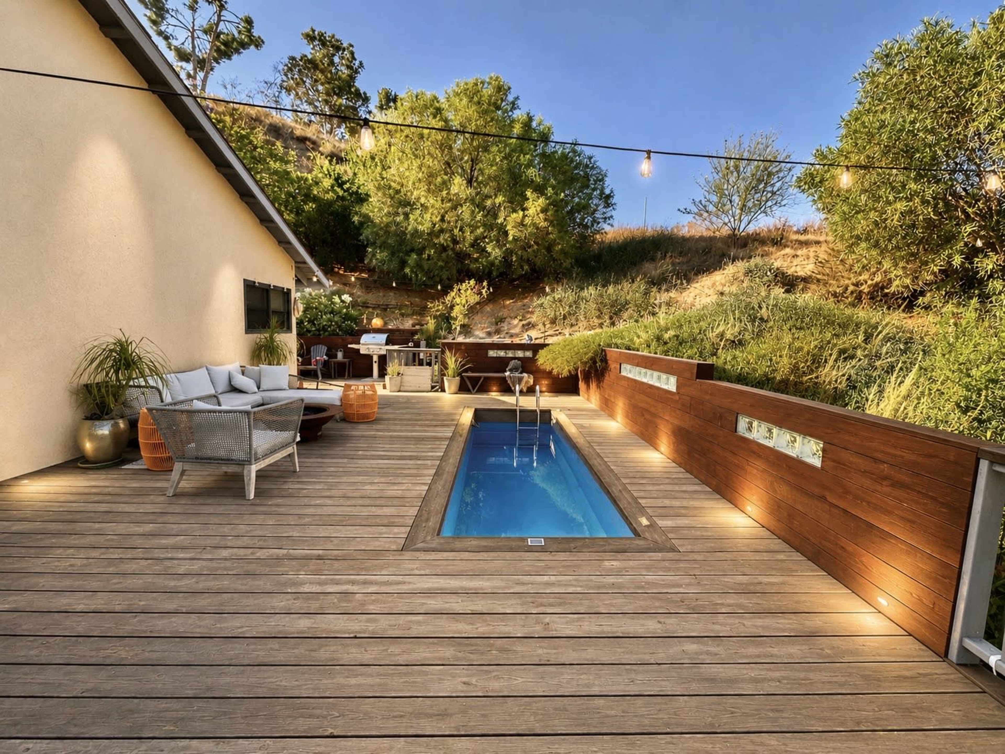 The image shows a wooden deck with a small swimming pool, surrounded by greenery and fairy lights hanging above.