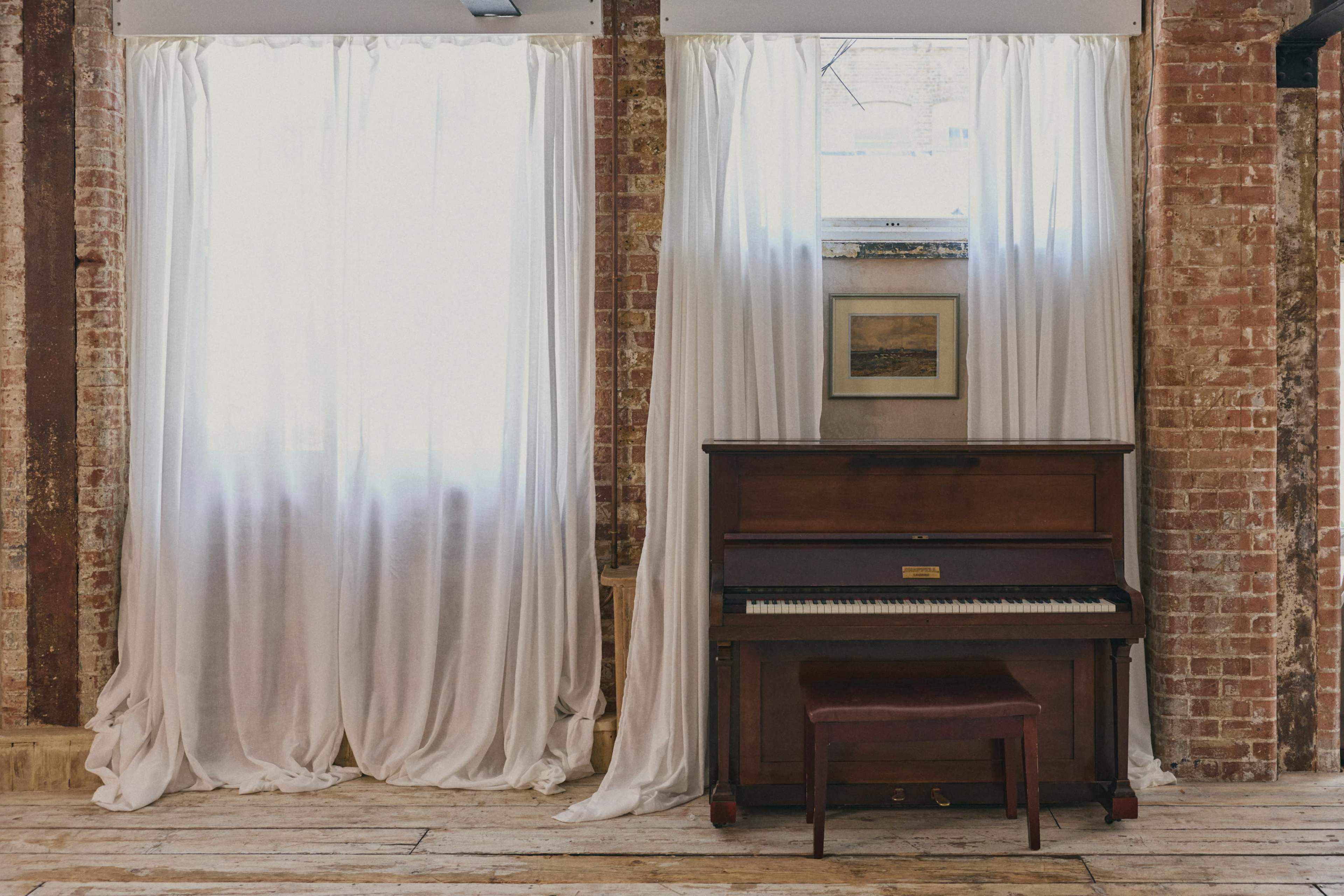 A wooden piano with a matching stool is positioned between two sheer white curtains and a framed picture on a brick wall.