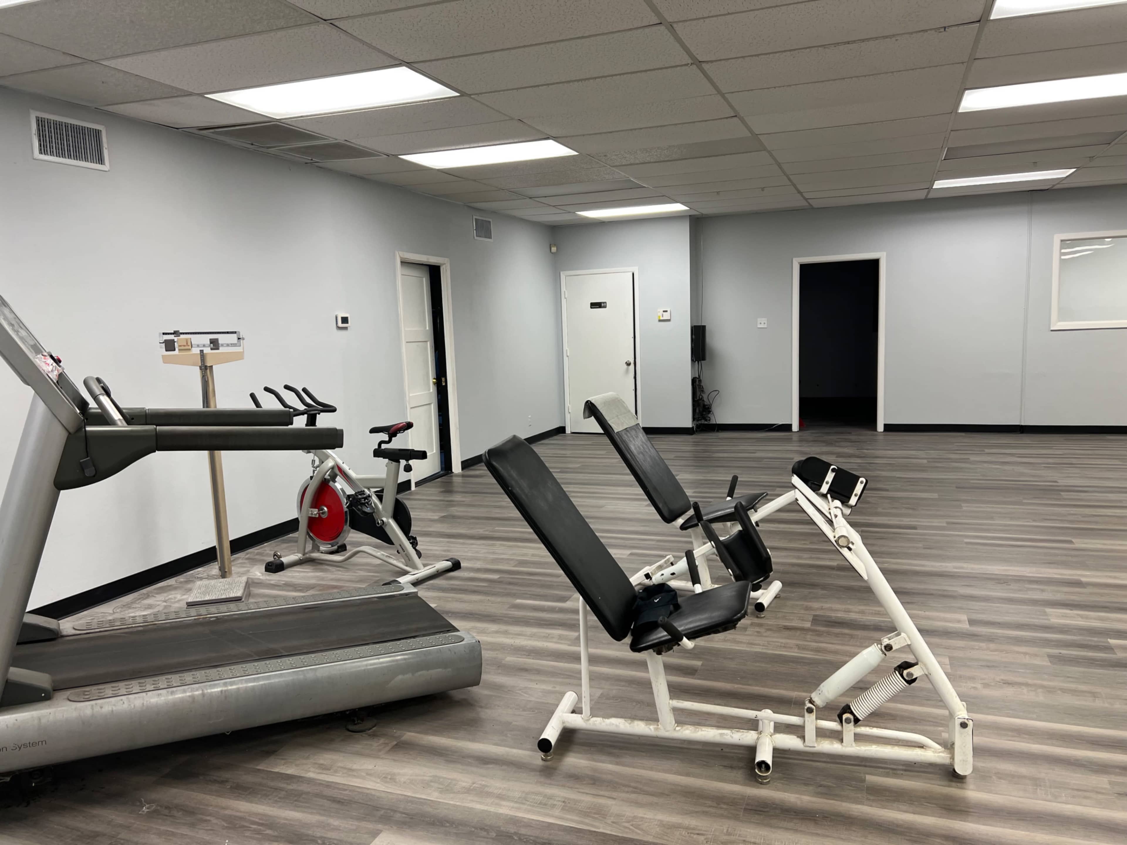 The image shows a sparsely equipped gym with a treadmill, a stationary bike, and a weight bench arranged in a well-lit room.