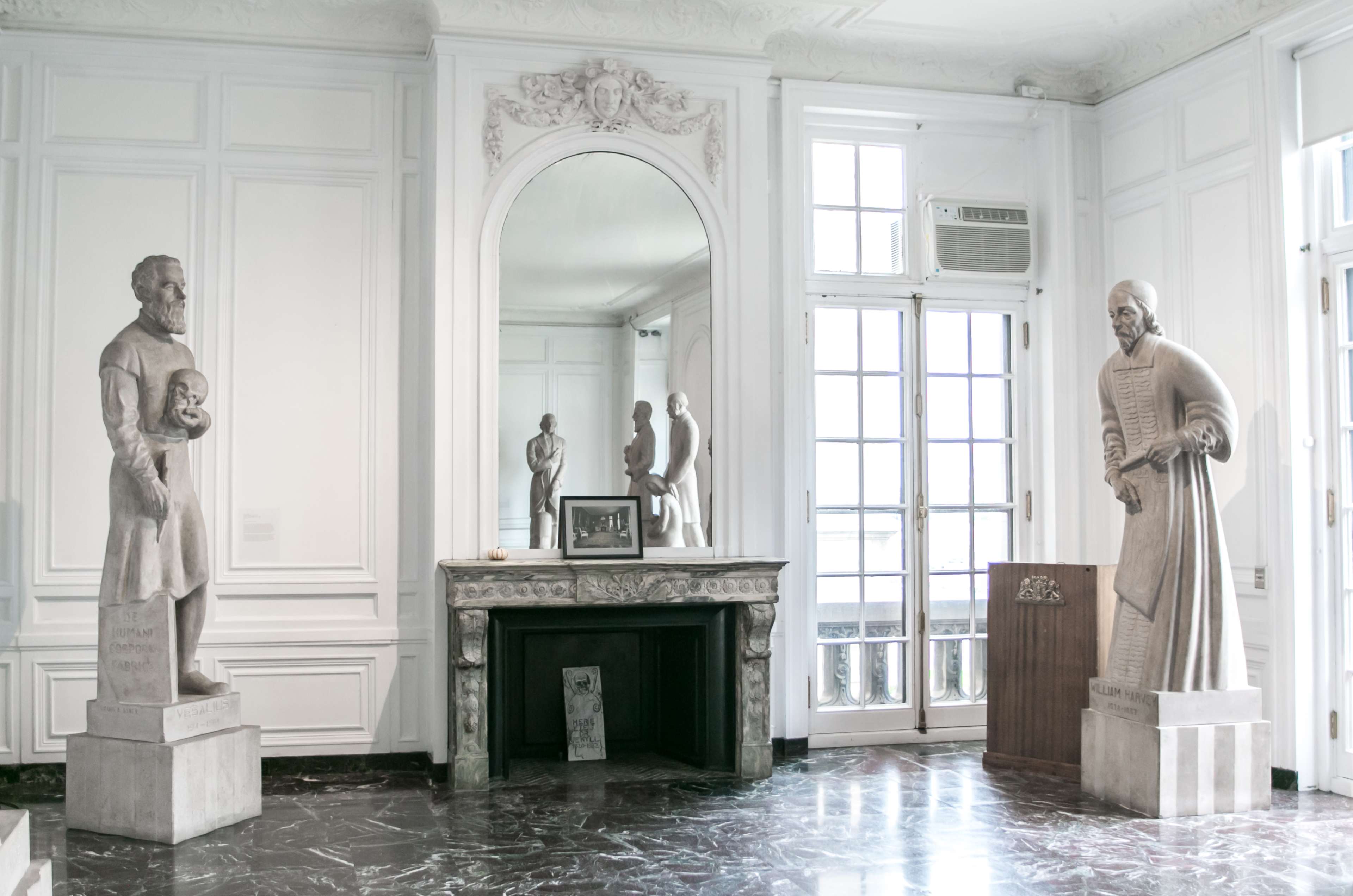 A bright room with two statues on pedestals facing each other, a fireplace with a decorative mantel, and large windows letting in natural light.