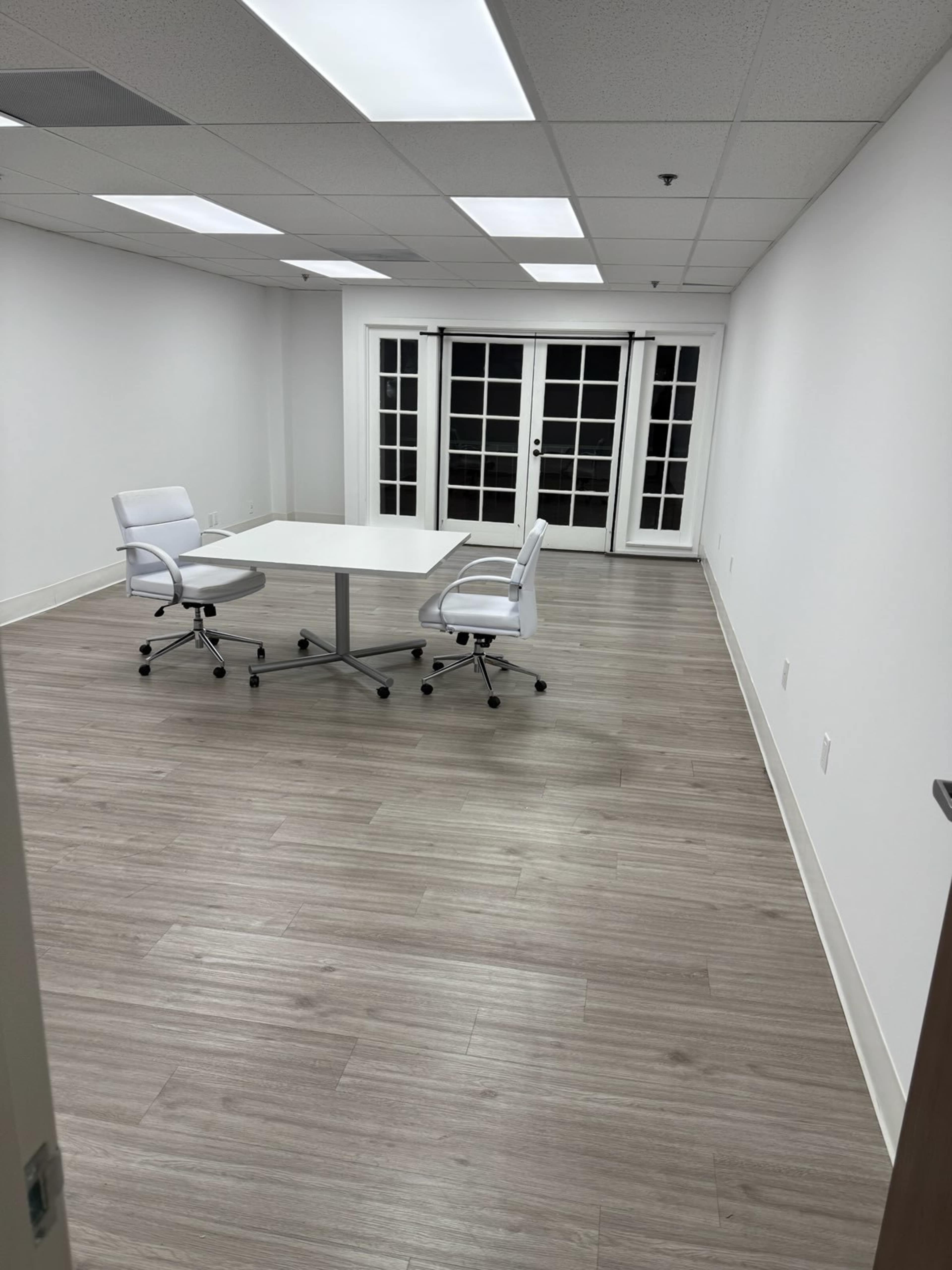 A sparse office meeting room features a square table and two white chairs, with a set of glass doors leading to another space.