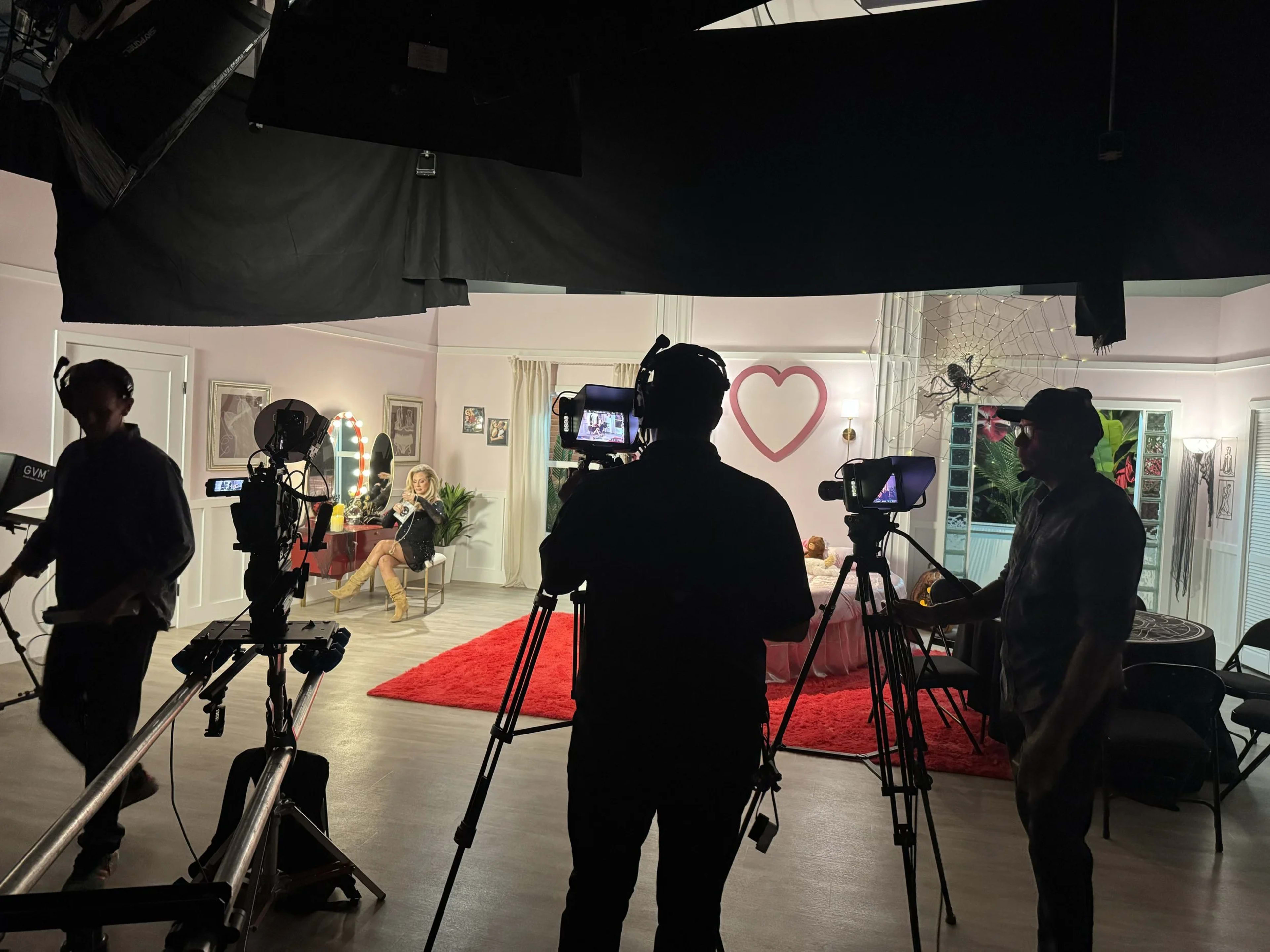The image shows a film set with three camera operators positioned in the foreground, filming a scene in a brightly lit room decorated with a large heart and soft furnishings.