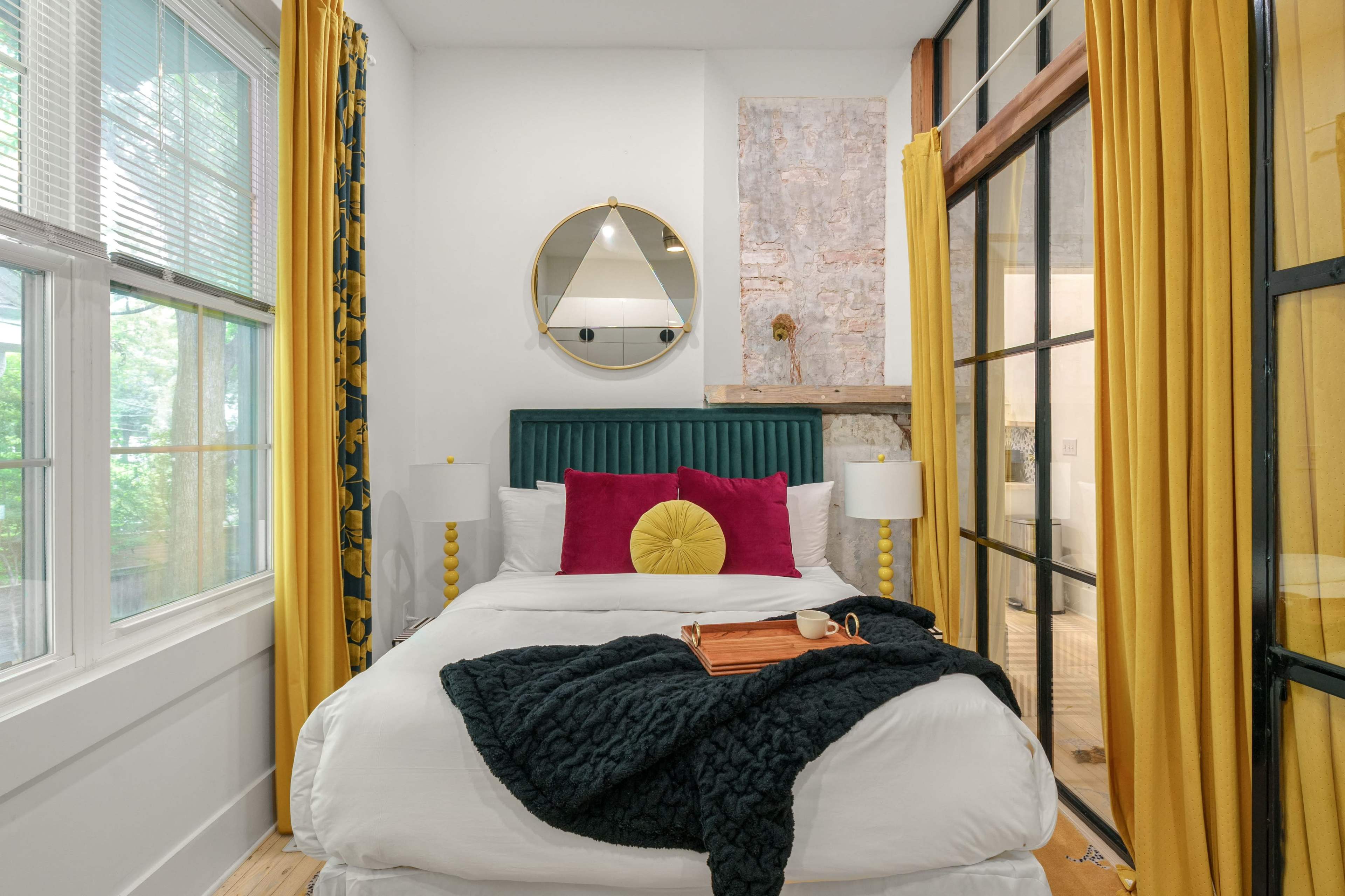 The image shows a well-decorated bedroom featuring a bed with a green headboard, colorful pillows, yellow curtains, and large windows allowing natural light to enter the space.