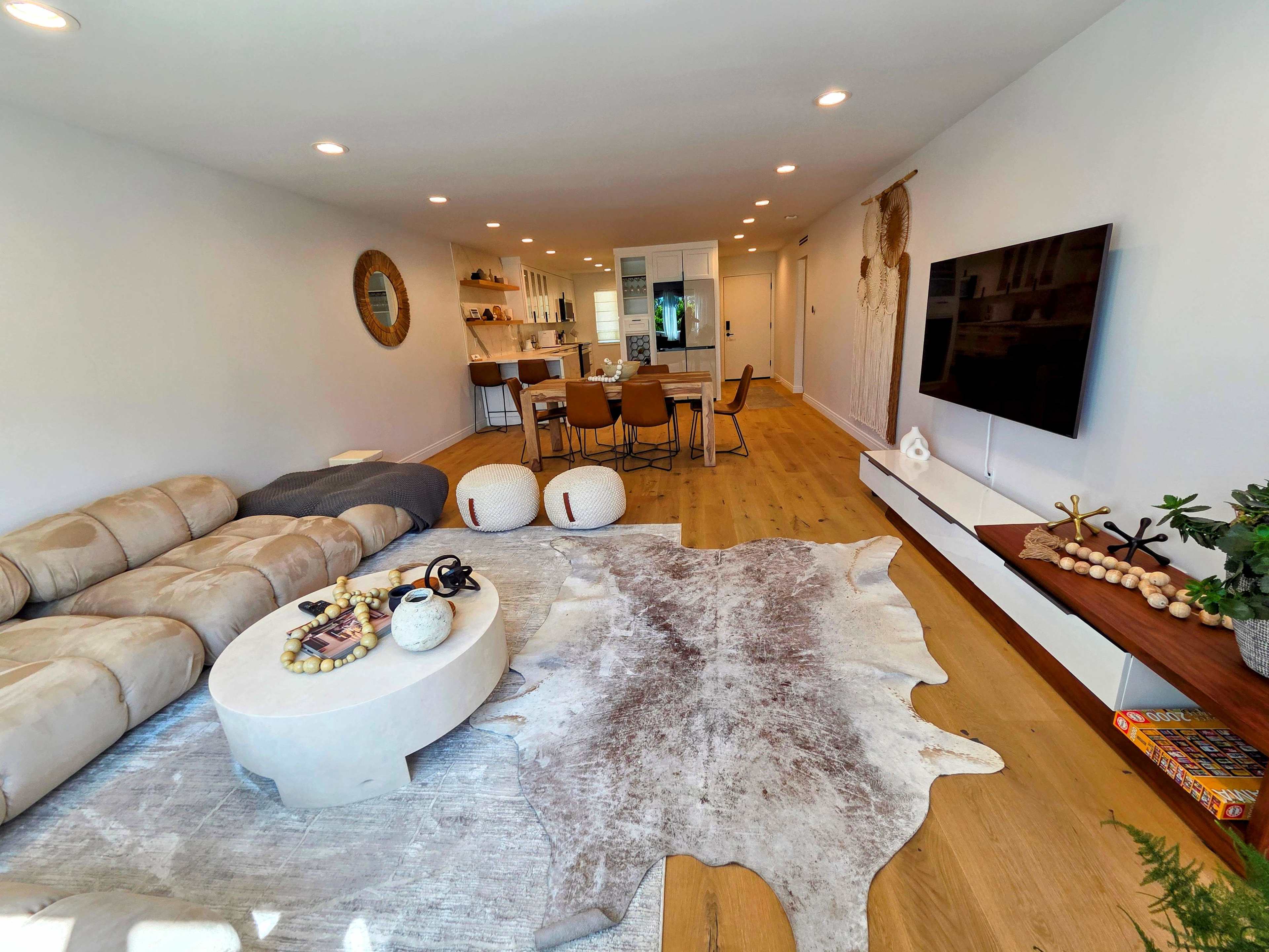 The image shows a modern living room with a large sectional sofa, a circular coffee table, a decorative rug, and a dining area in the background featuring a wooden table and chairs.
