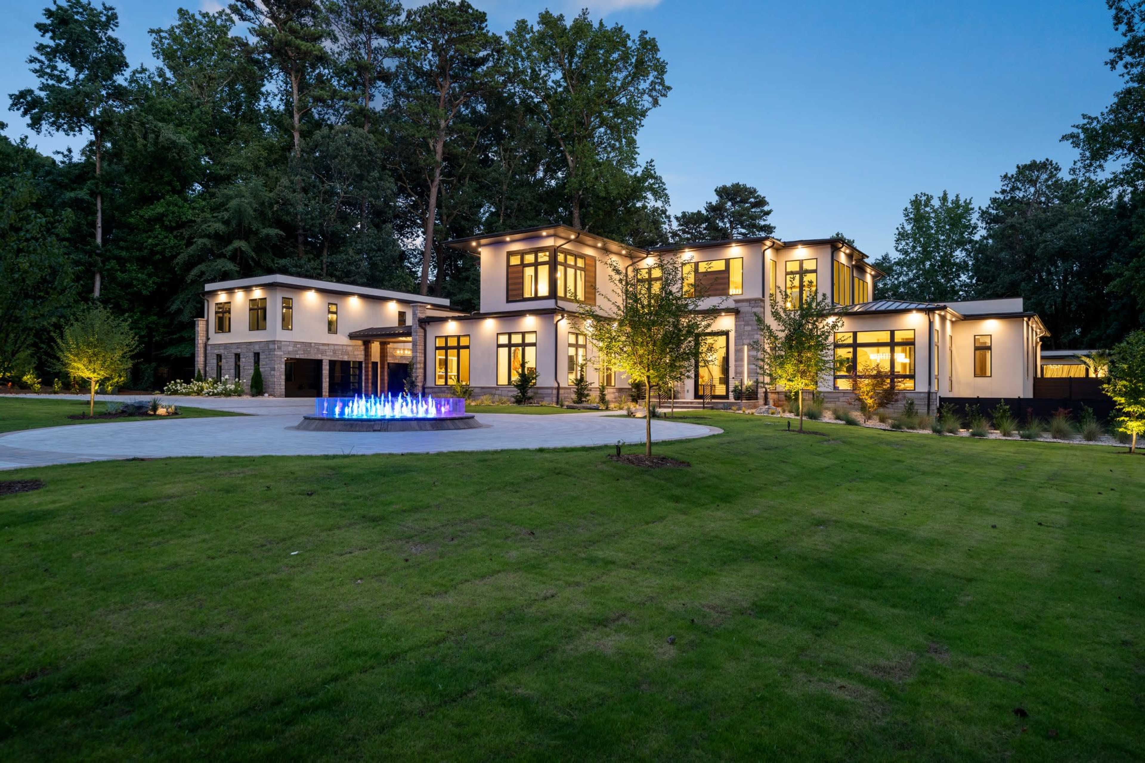 A modern two-story home with large windows and stone accents is surrounded by a landscaped yard and features a circular water fountain with illuminated jets.