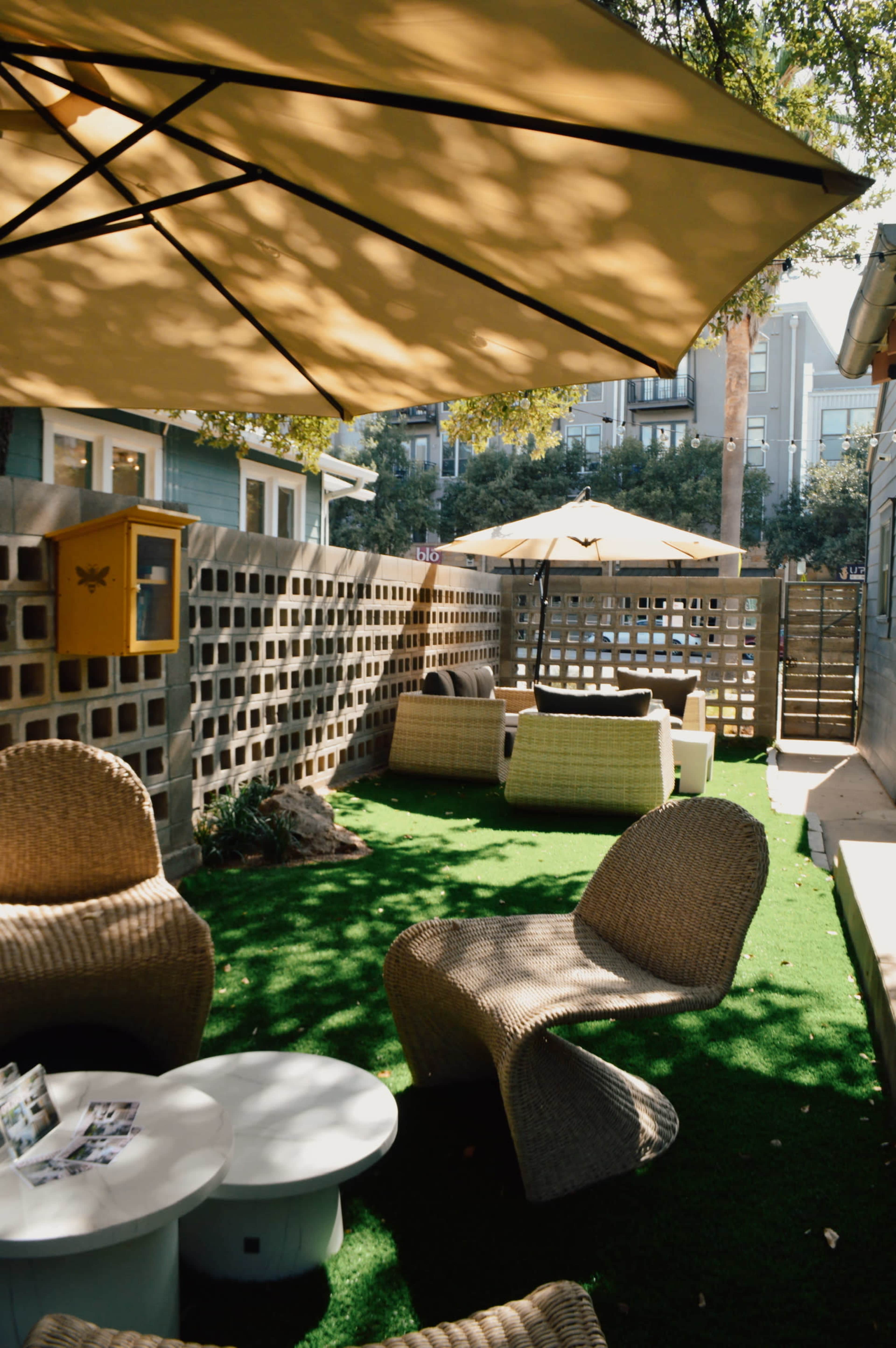 The image shows a landscaped outdoor area with seating made of woven material, shaded by large umbrellas and surrounded by a decorative wall.