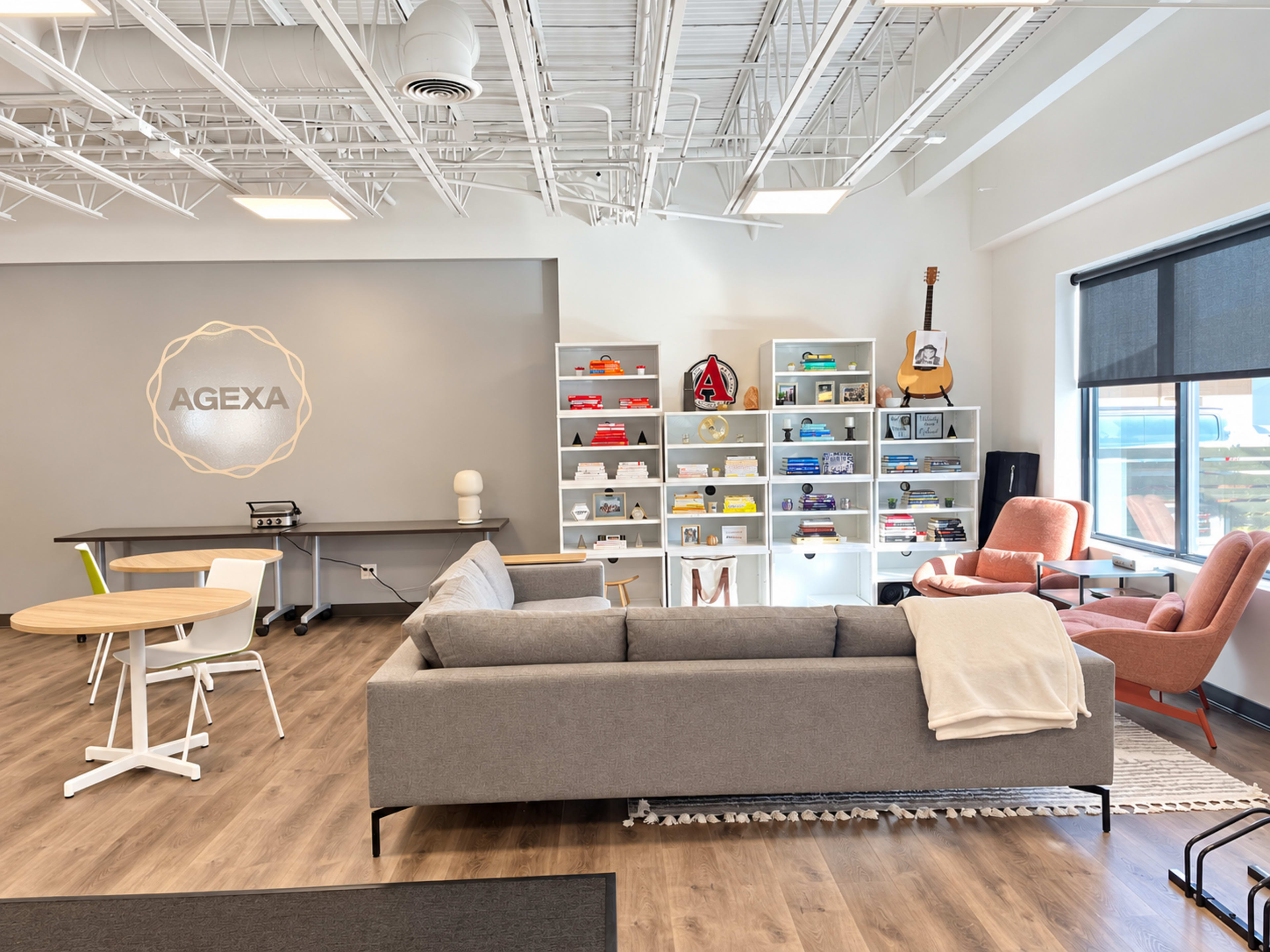 The image shows a modern office lounge area with a grey sofa, a round wooden table, colorful shelves filled with items, and a guitar leaning against a wall.