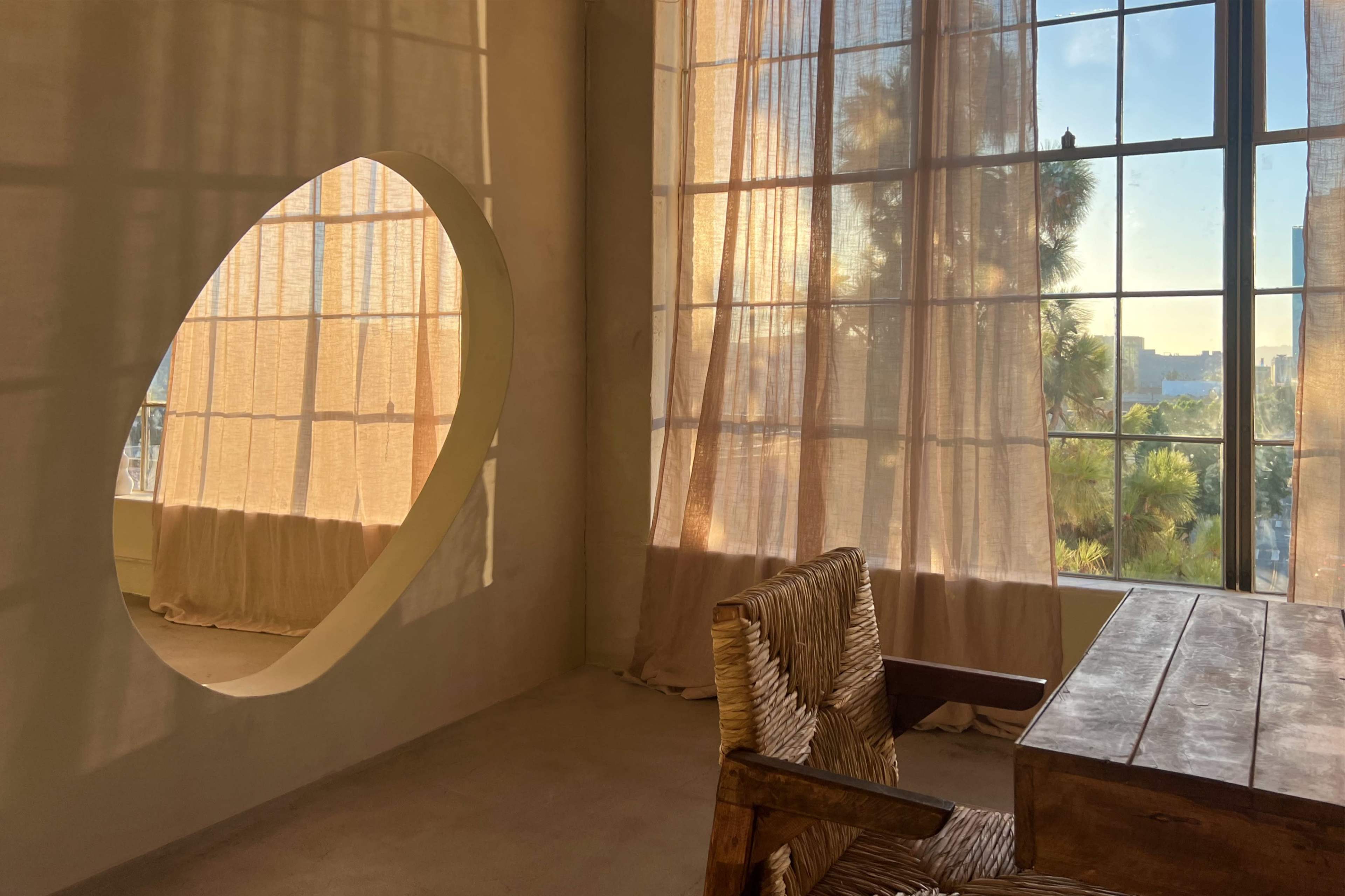 A room with large windows covered by sheer curtains and a round cutout reflecting light, featuring a wooden desk and woven chair.
