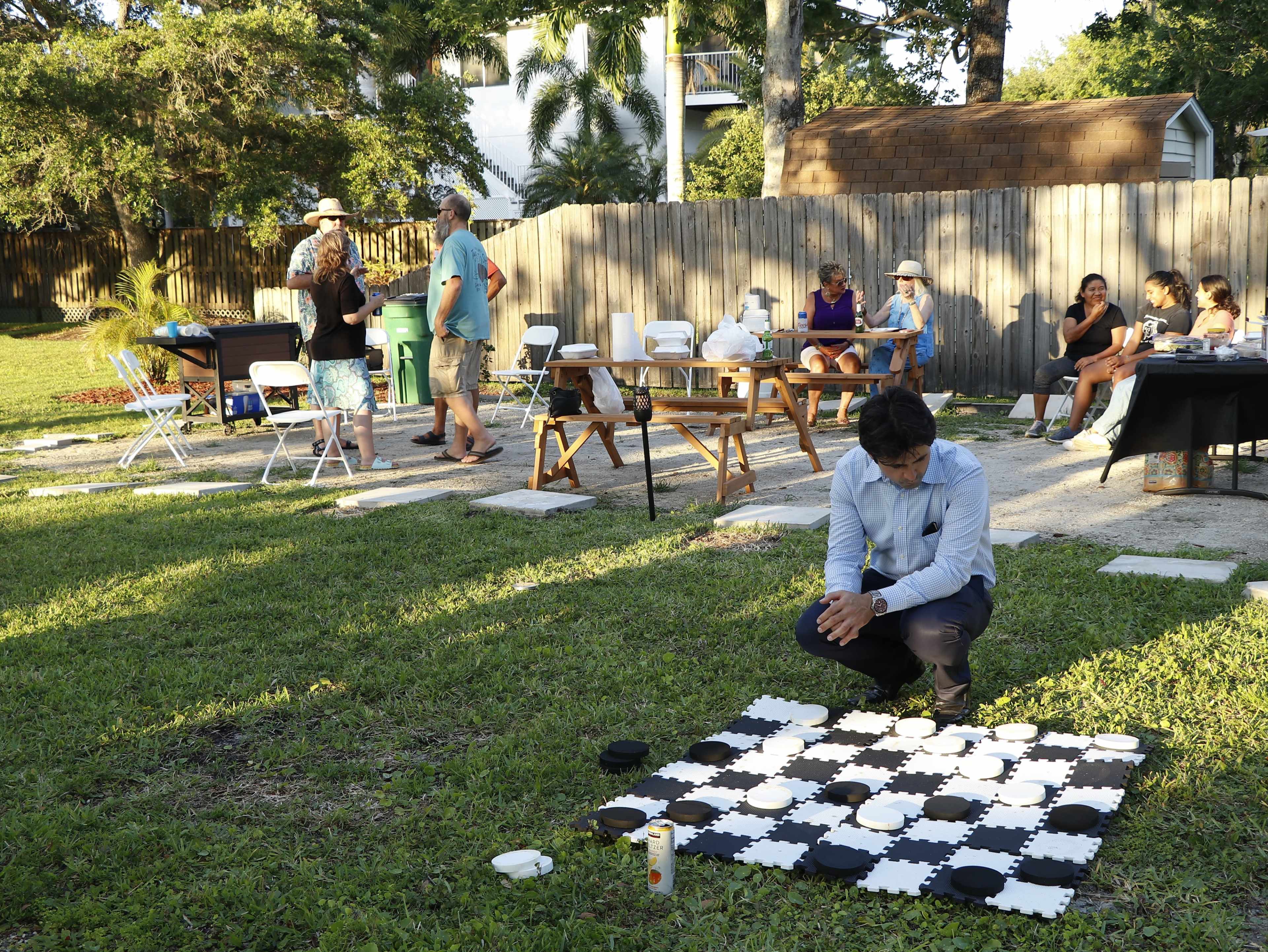 A group of people socializes in a park while a young man plays a game on a large checkers board on the grass.