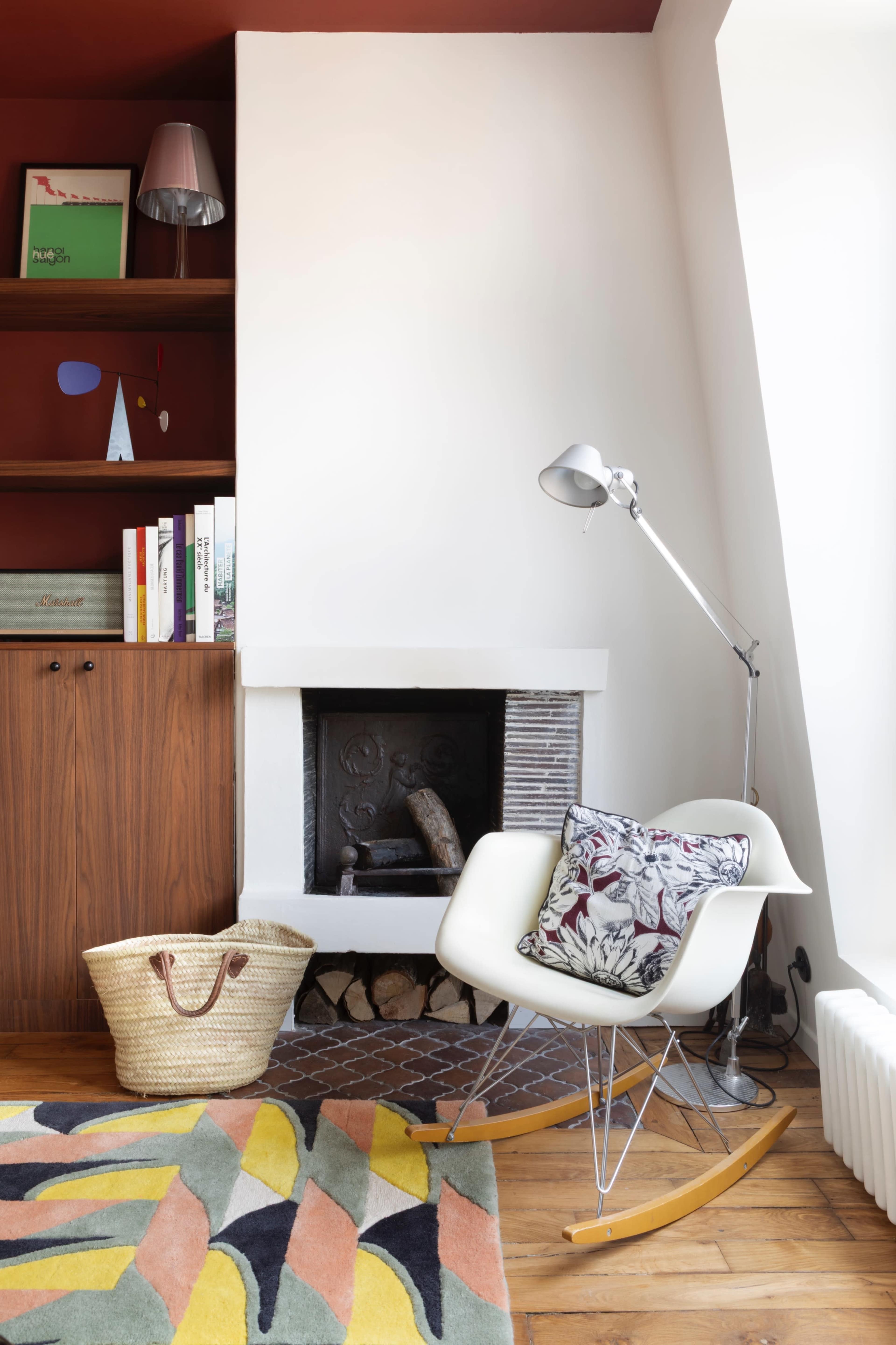A white rocking chair with a decorative pillow is positioned beside a modern floor lamp and a built-in bookshelf in a room with a red accent wall.