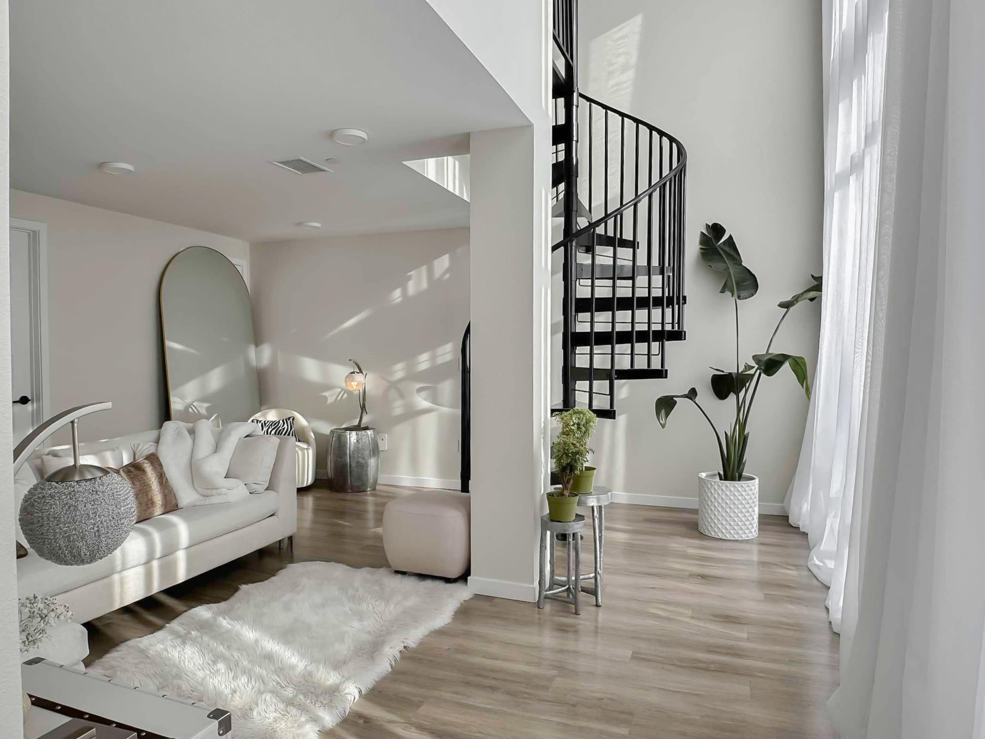 The image shows a modern living space featuring a spiral staircase, a light-colored sofa, a plush rug, and several potted plants in a well-lit room.