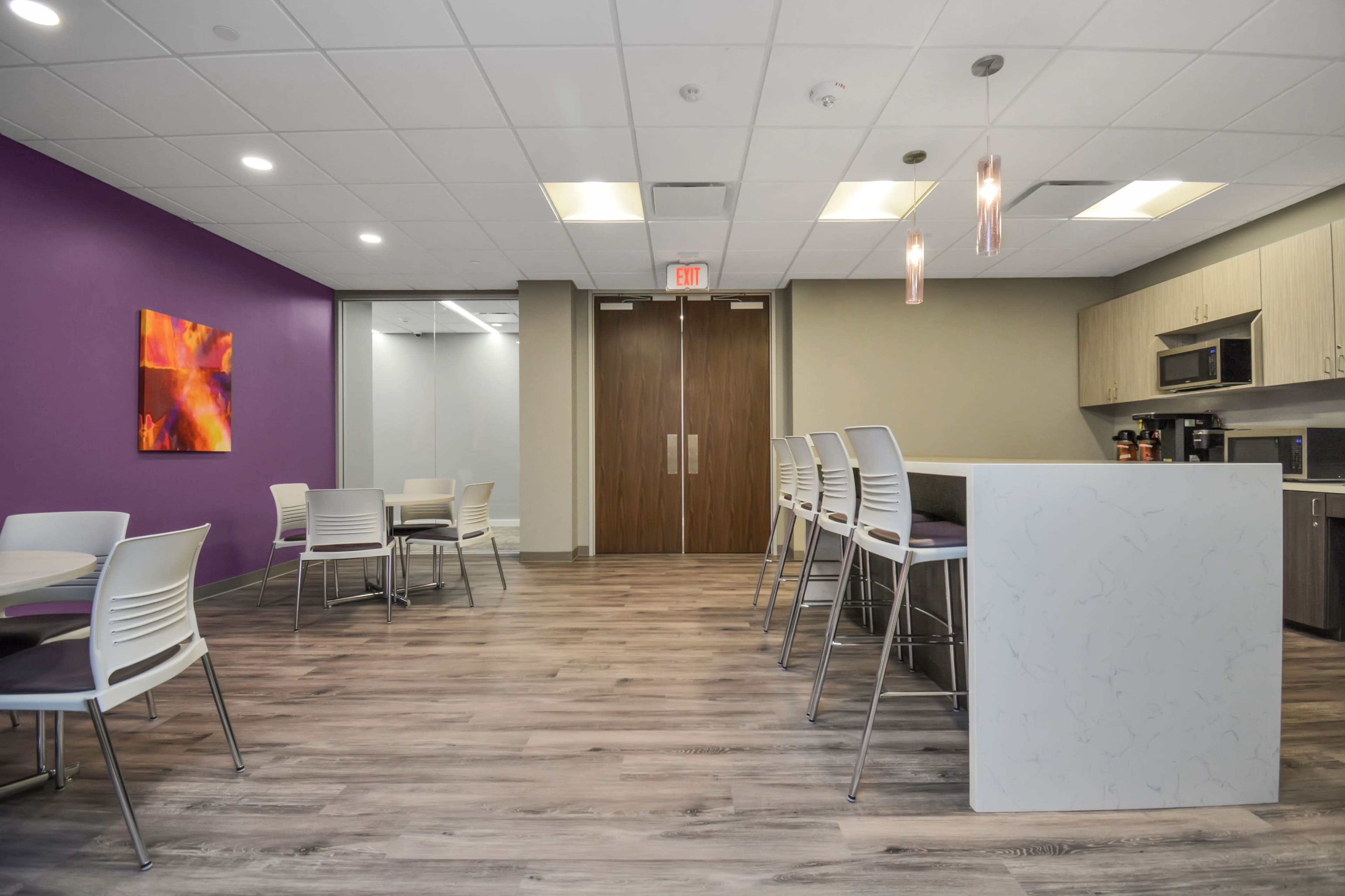 The image shows a modern break room with a purple accent wall, a kitchenette area, and several white tables and chairs.