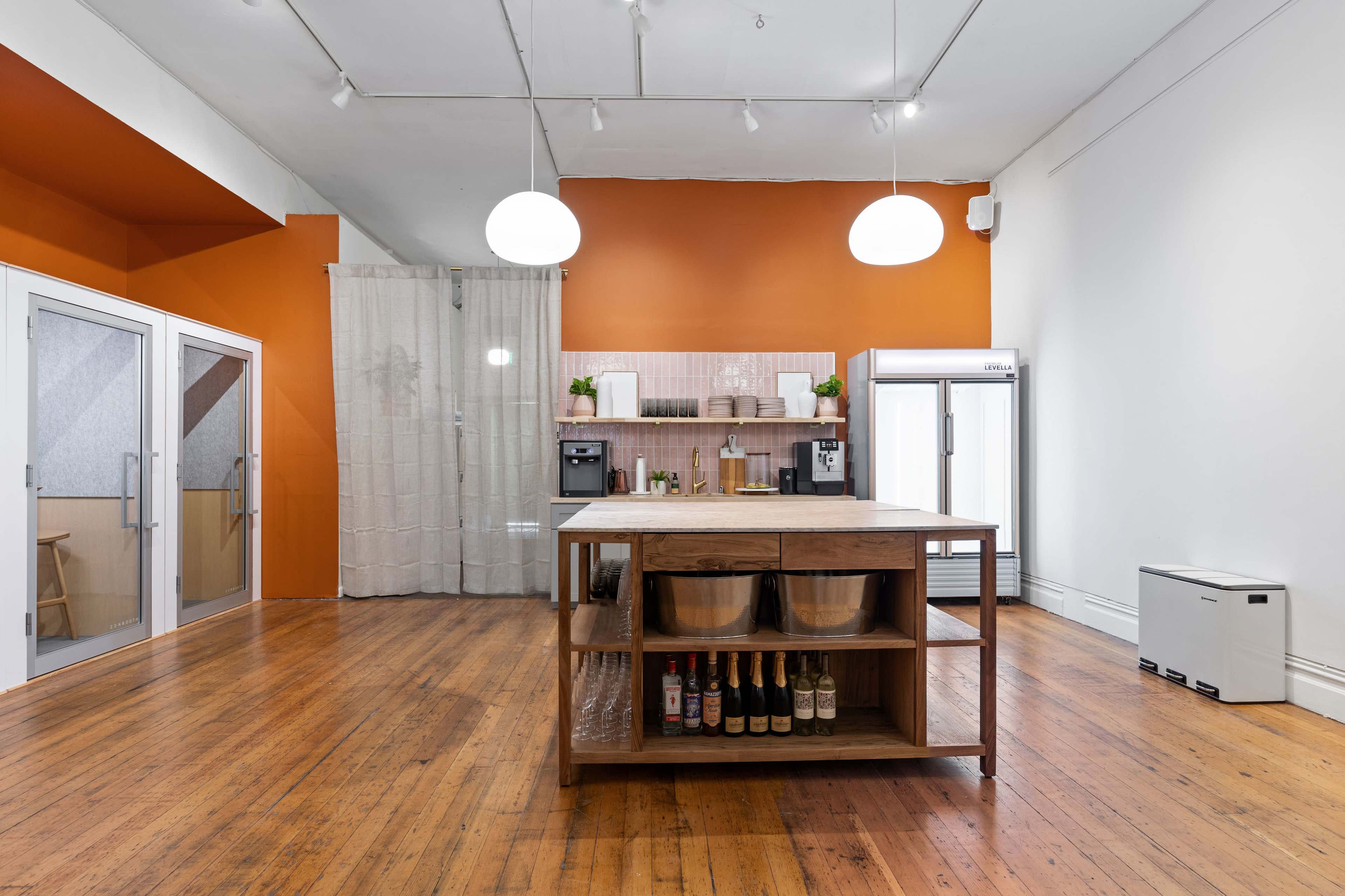 A spacious room with wooden floors featuring a central wooden island, a kitchen area with modern appliances, and an orange accent wall.