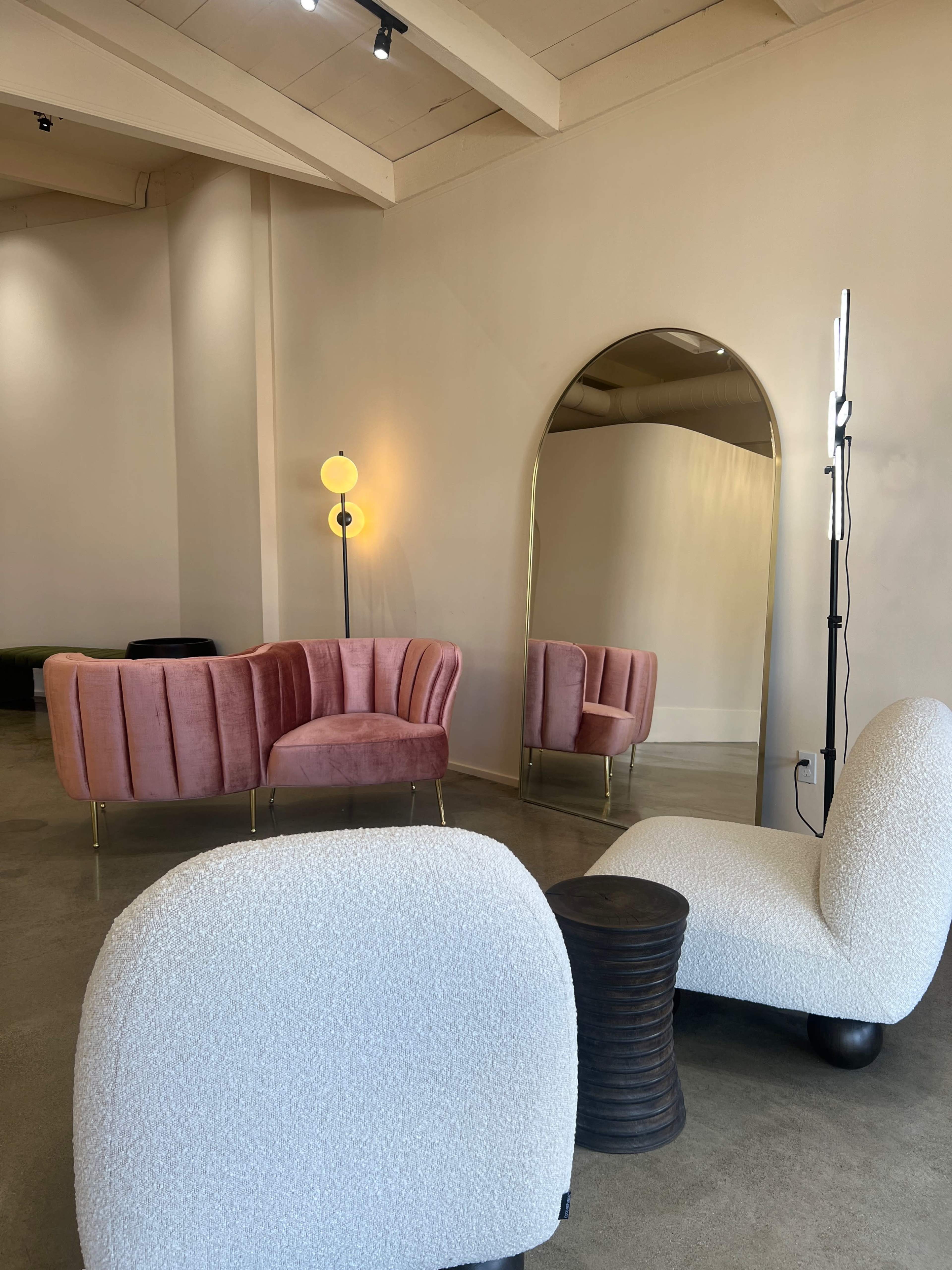 A modern interior featuring a pink velvet sofa, a large mirror, a sculptural chair, and a small black side table.