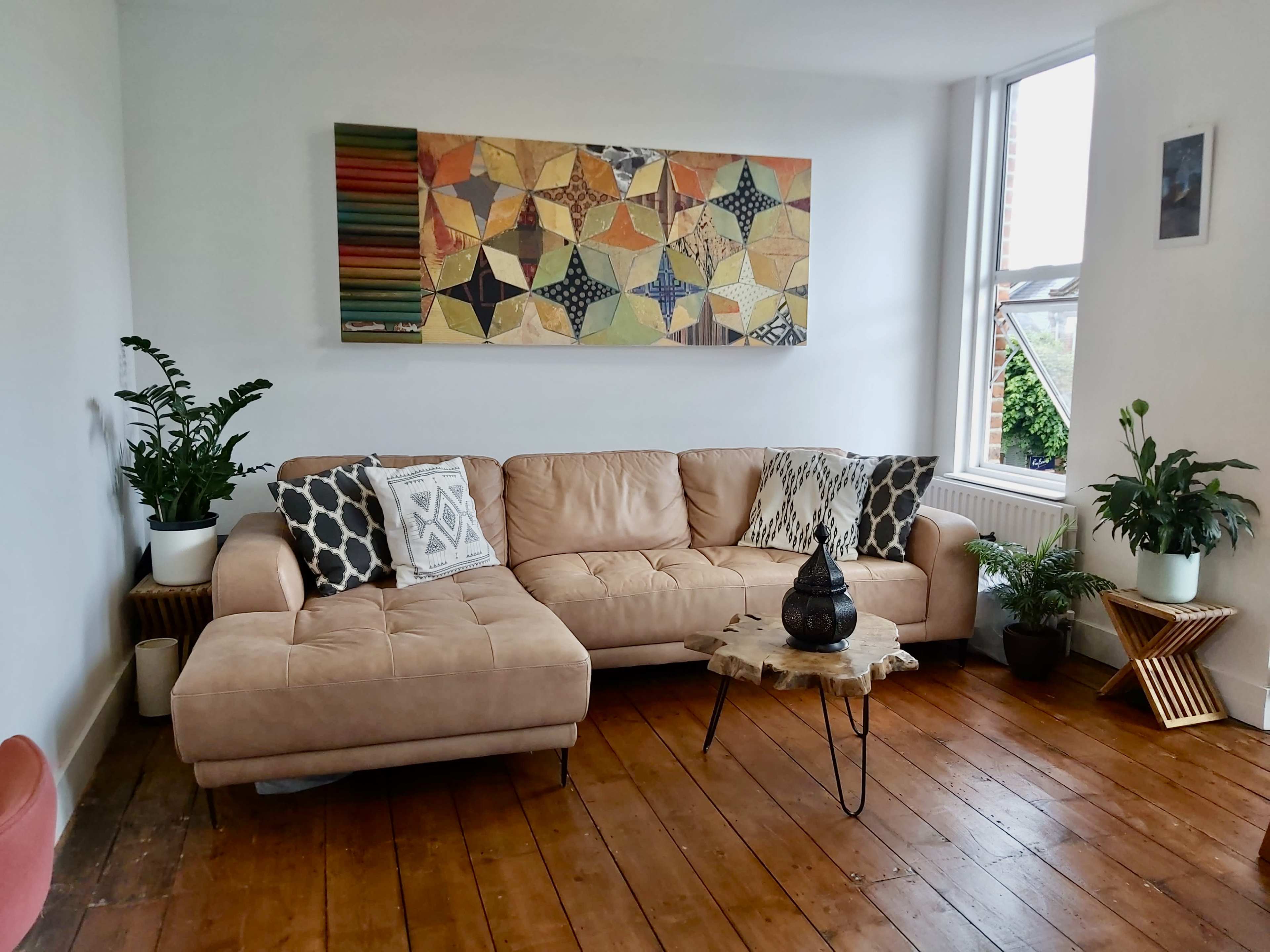 A cozy living room with a light-colored sectional sofa, a colorful wall art piece above it, and various plants in decorative pots.