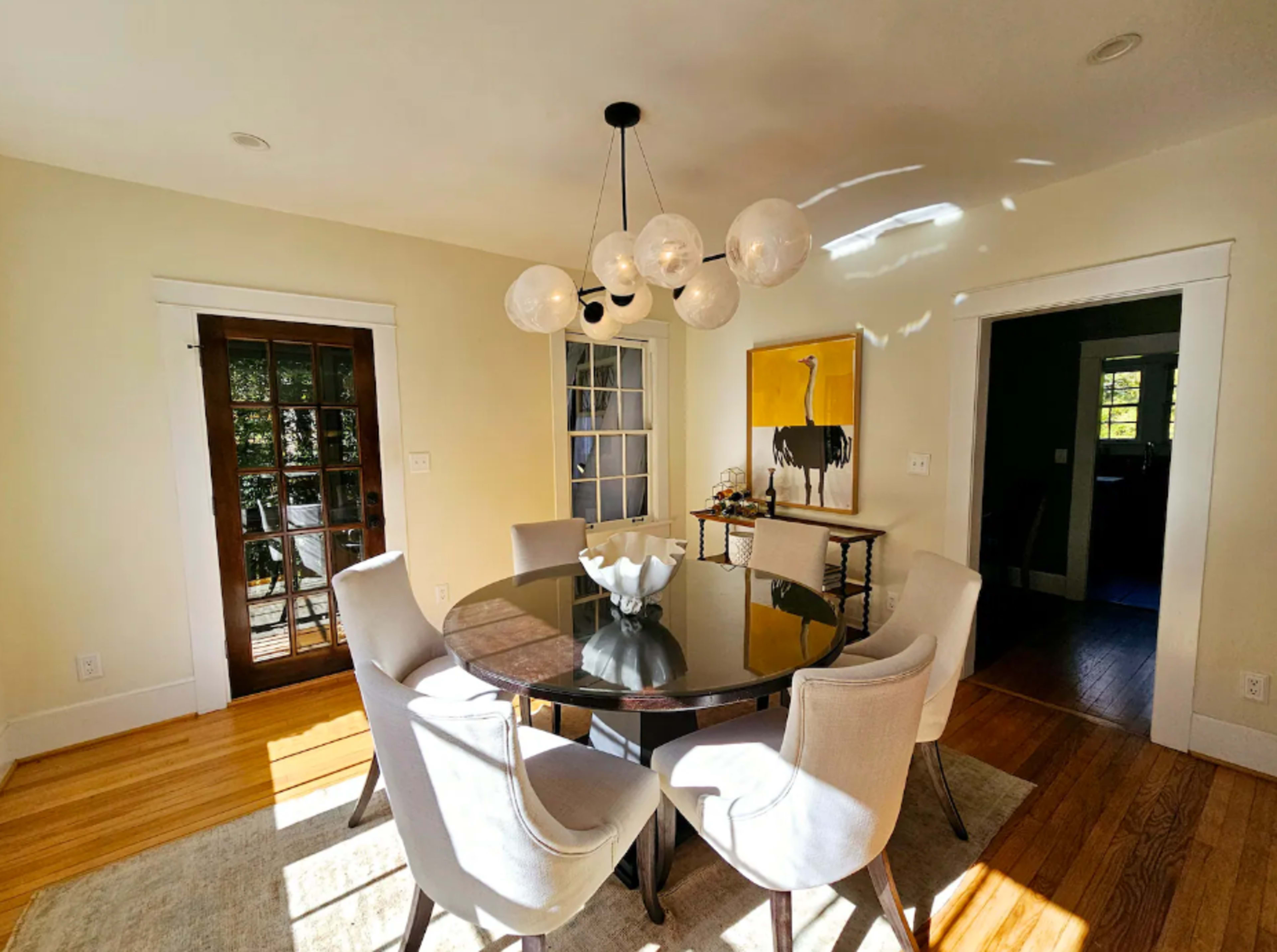 The image shows a dining area with a circular black table surrounded by six light-colored chairs, illuminated by a cluster of pendant lights, with a painting of an ostrich on the wall.