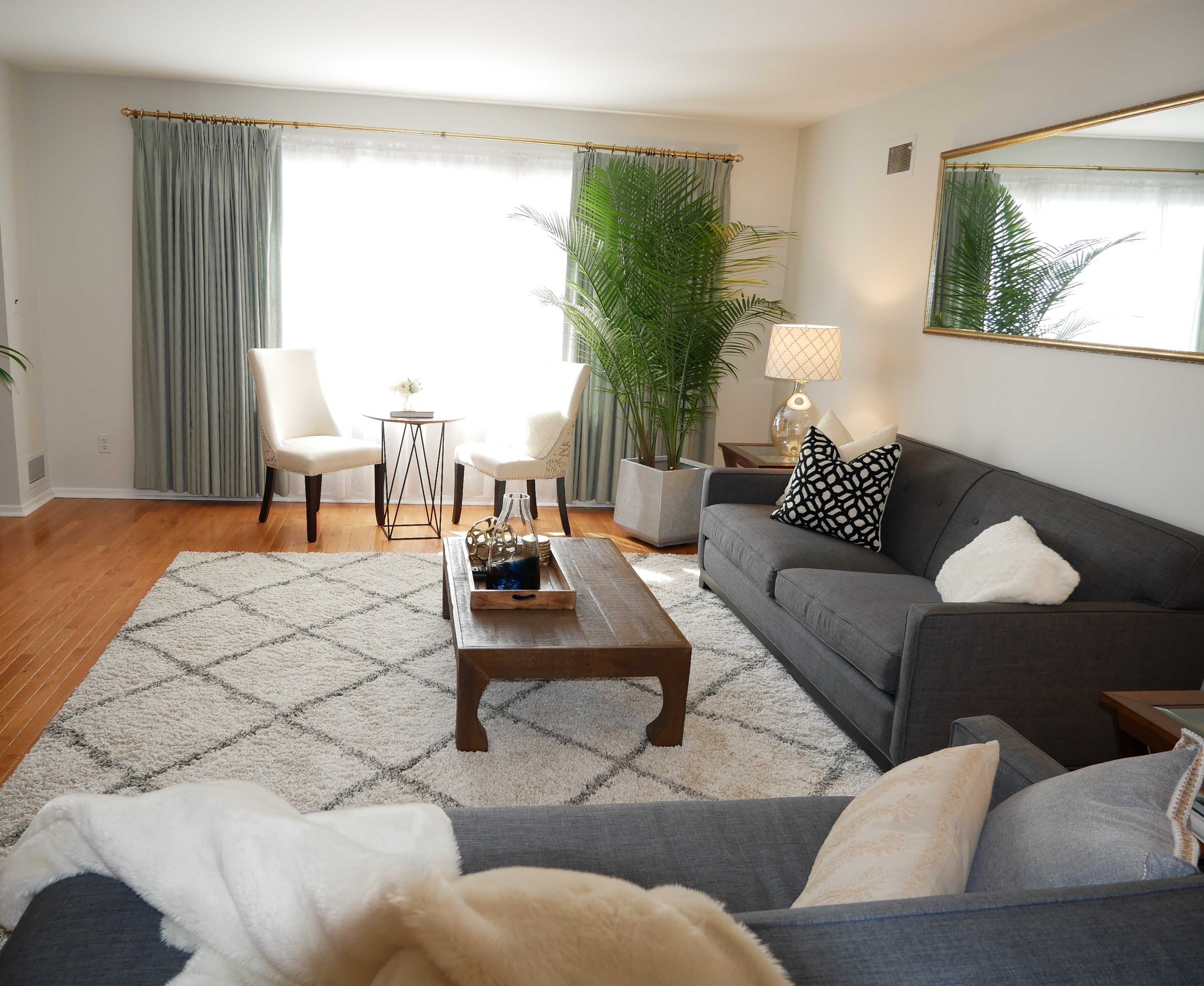 The living room features a gray sofa, a wooden coffee table, two chairs near a small side table, large potted plants, and large windows with sheer curtains.