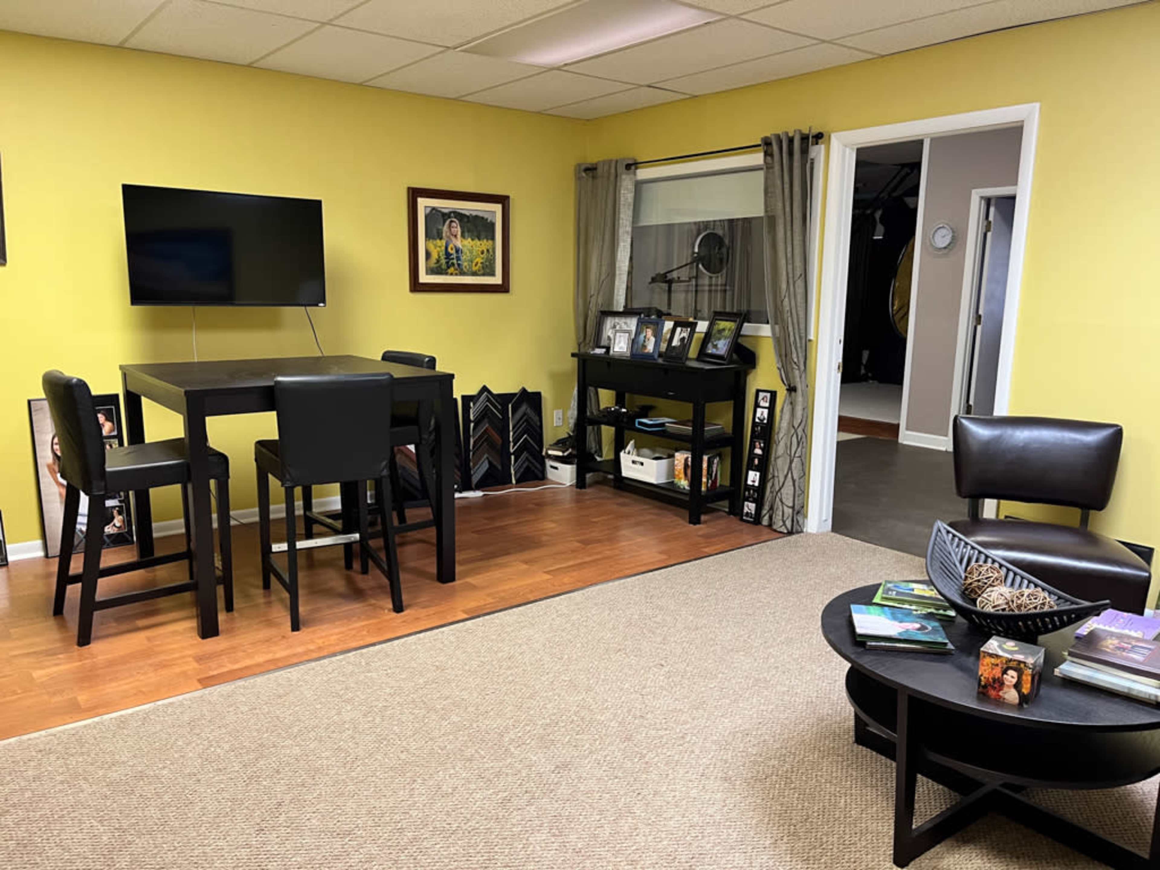 A brightly lit room with yellow walls, featuring a black dining table and chairs, a wall-mounted television, and a coffee table with books.