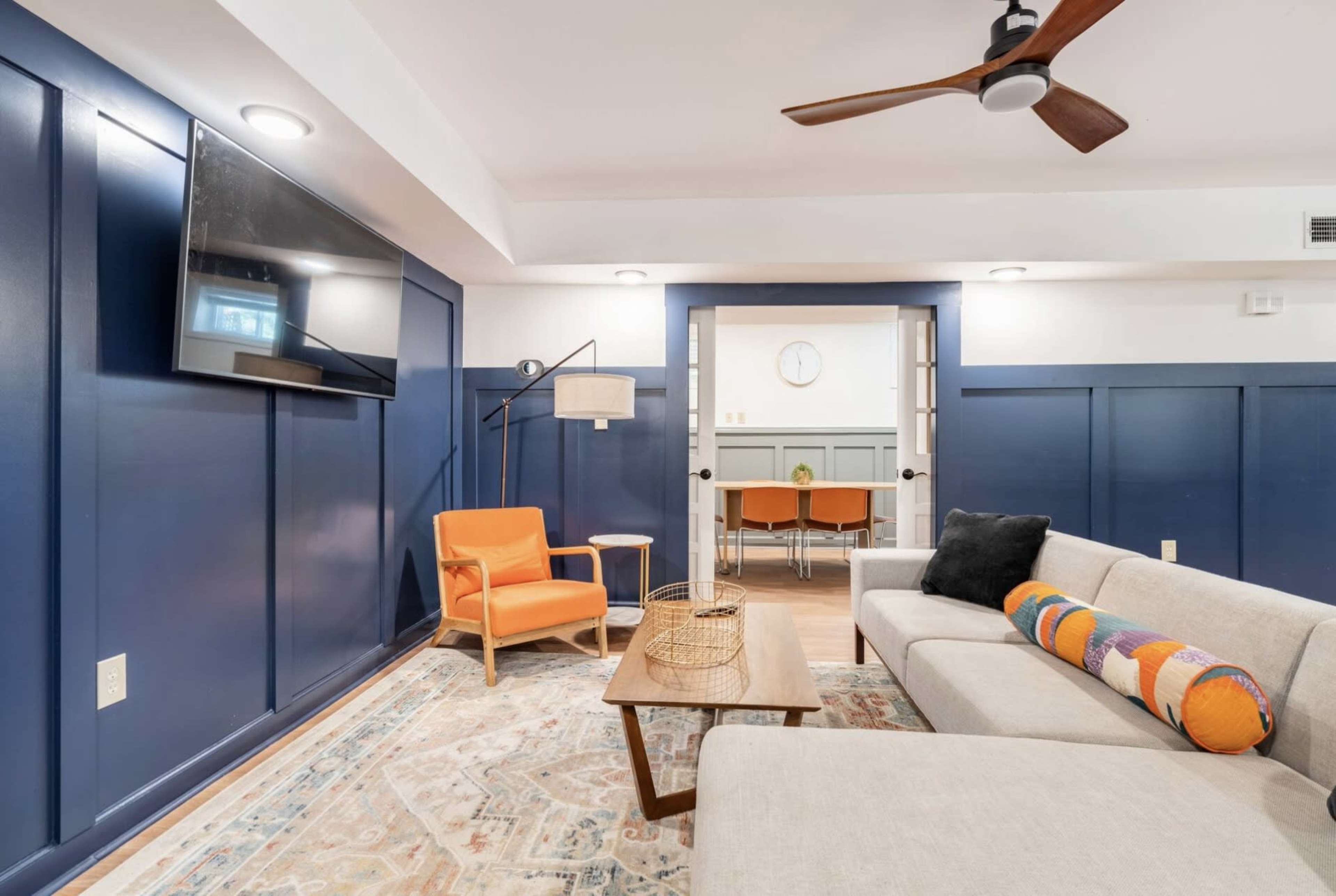 A living room with a sectional sofa, an orange armchair, a wooden coffee table, a flat-screen TV mounted on the wall, and a door leading to a dining area.