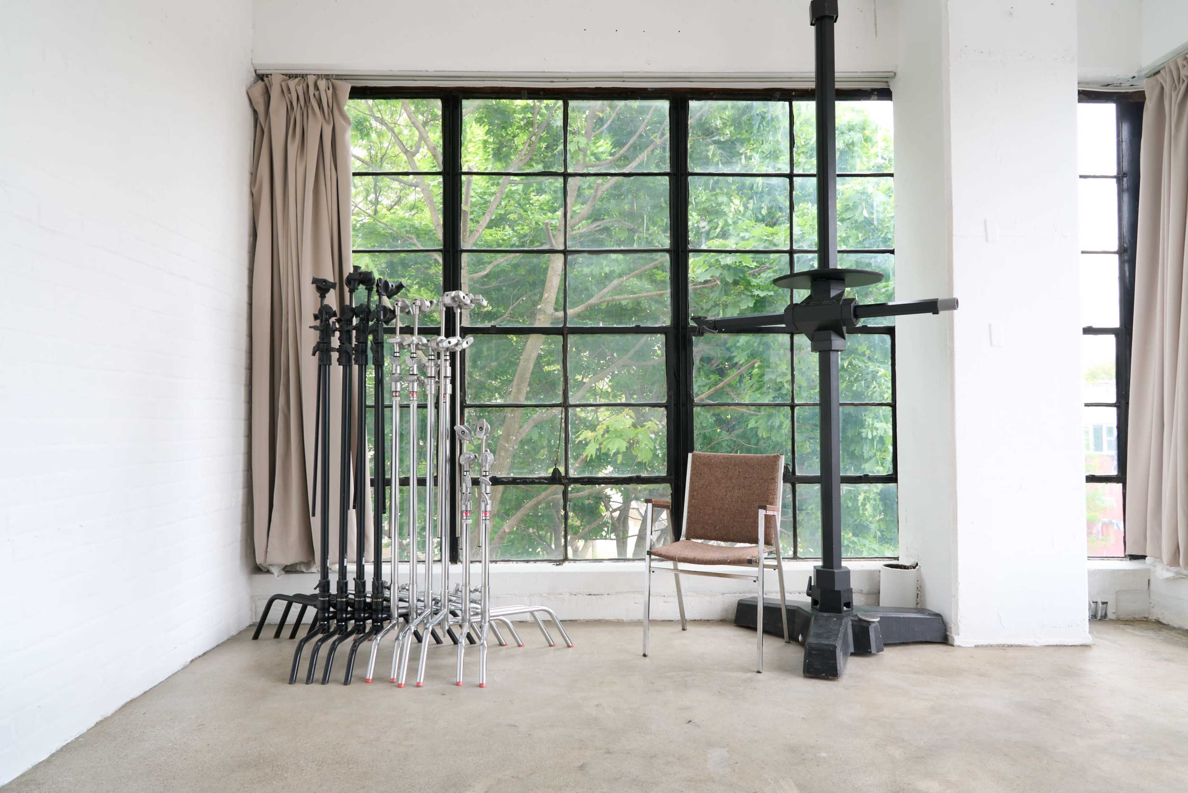 The image shows a minimalist interior with several lighting stands and a single chair beside large windows that overlook greenery outside.
