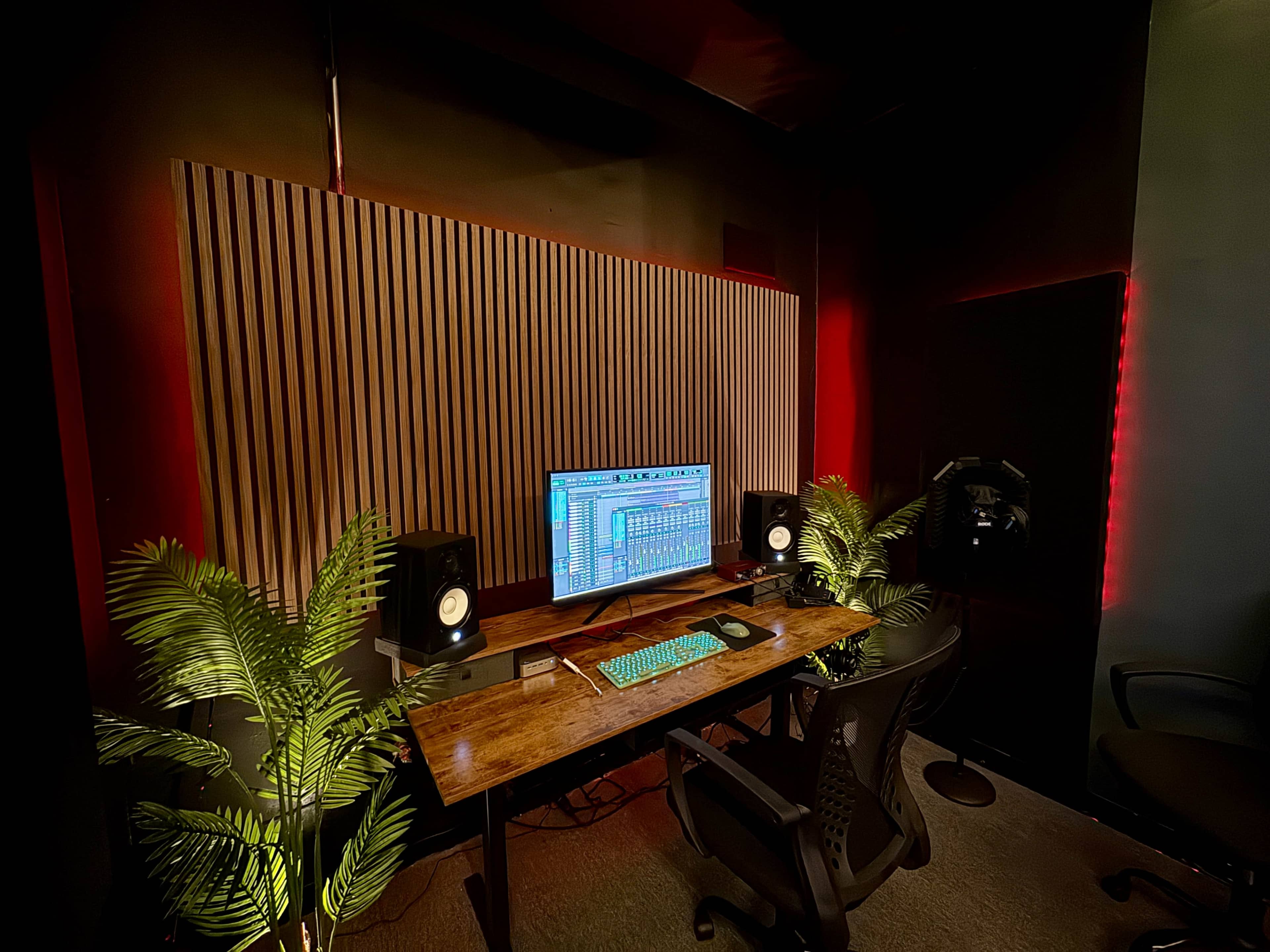 A music production studio features a wooden desk with a computer and speakers, surrounded by potted plants and illuminated by red lighting.