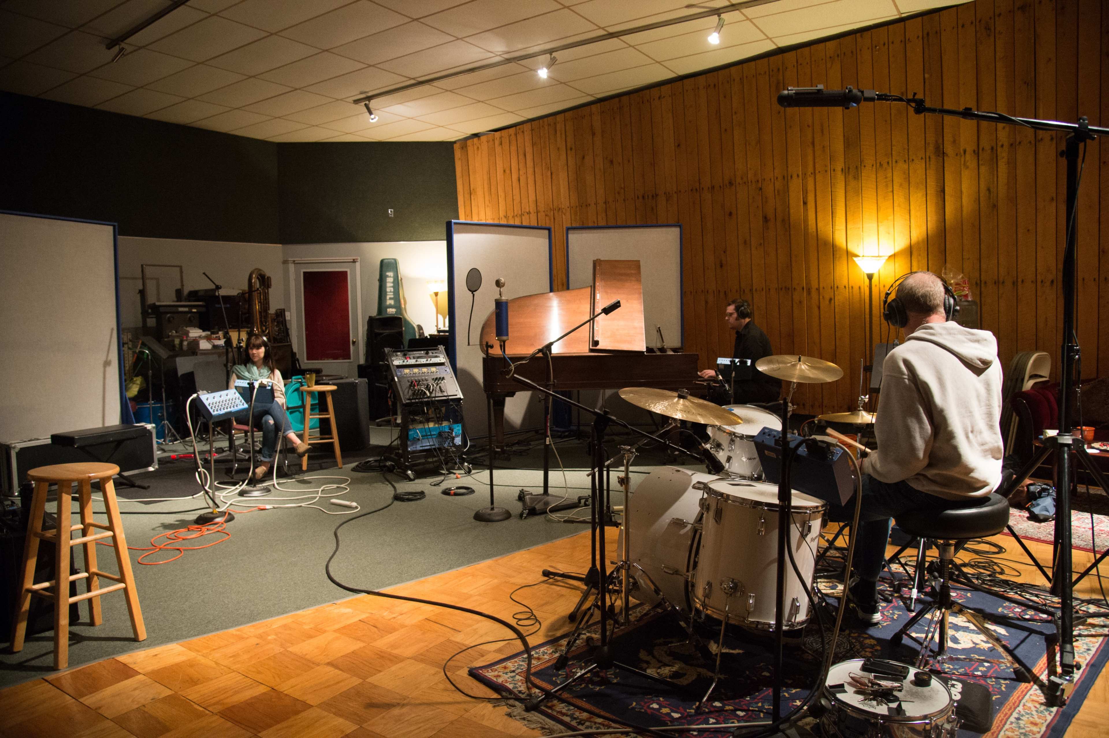 A recording studio features a drummer seated at a drum set, while two musicians are positioned at a keyboard and a piano in separate areas of the room.