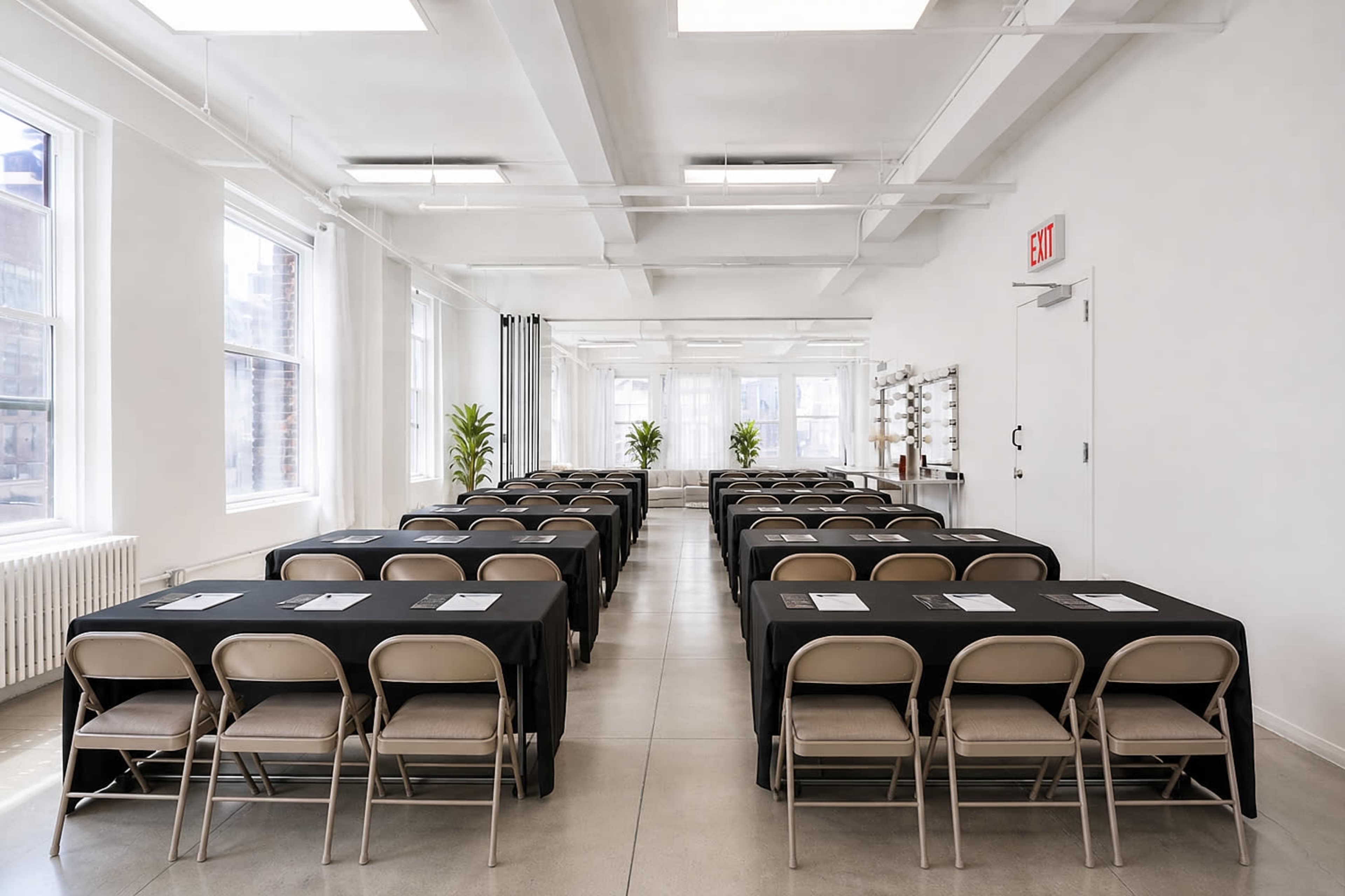The image shows a spacious meeting room with several rows of tables covered in black cloth and lined with chairs, flanked by large windows providing natural light.