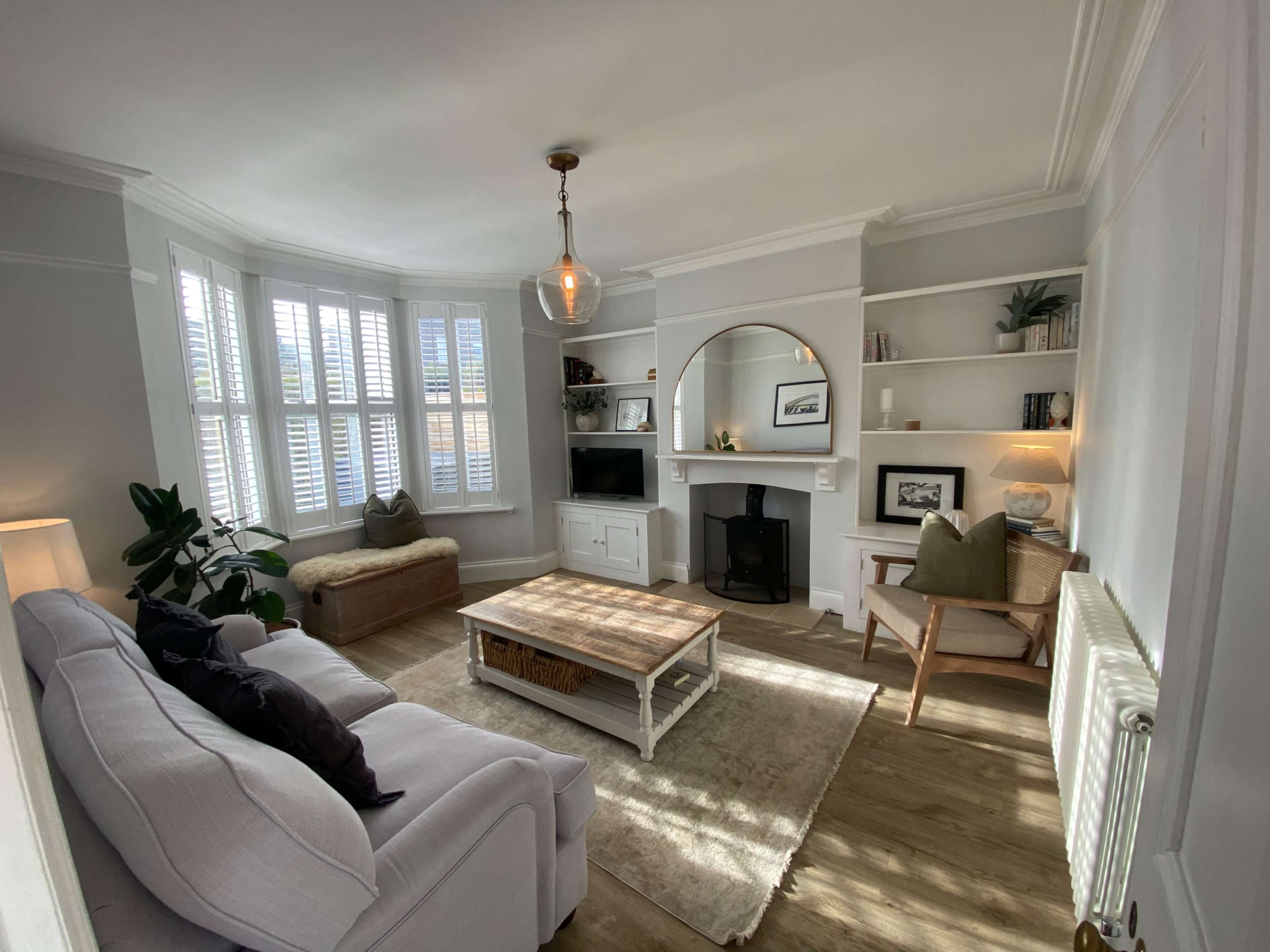 The image shows a bright living room with a bay window, a white sofa, a wooden coffee table, and shelves containing books and decorative items.