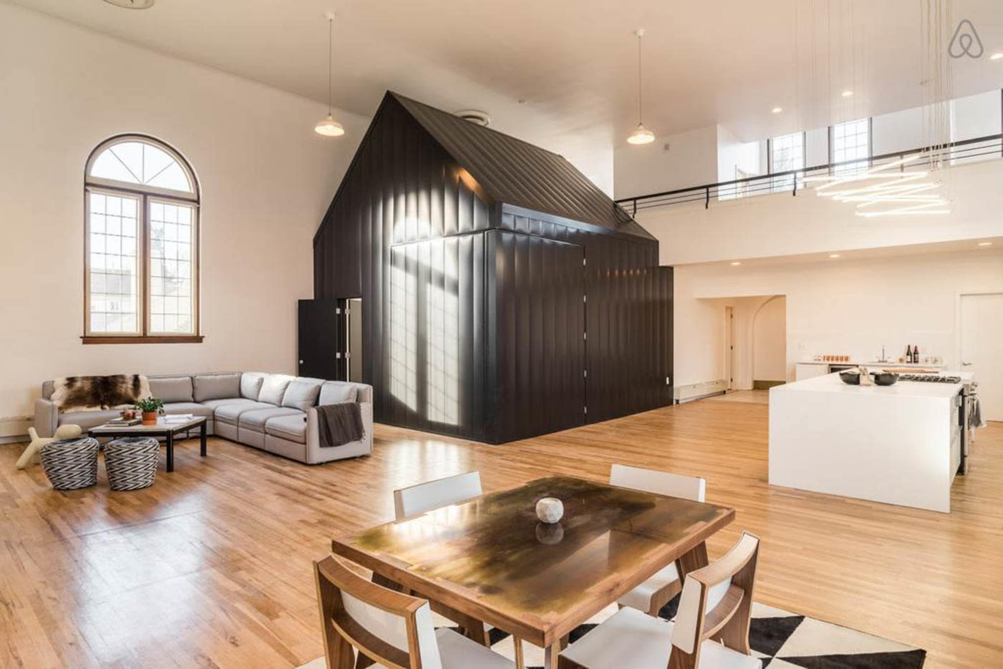 An open-concept living space featuring a modern kitchen, a dining area with a wooden table, and a black metal structure resembling a barn in the center.