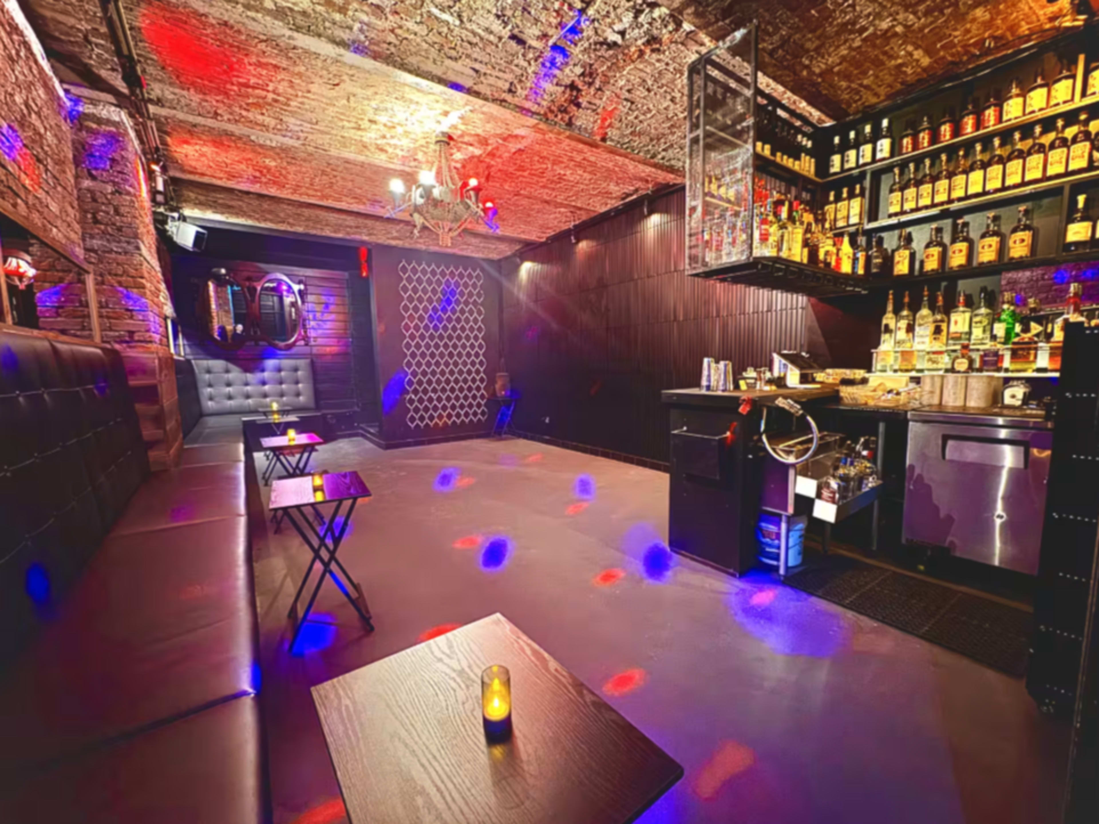 The image shows a dimly lit bar interior with a stone ceiling, shelves lined with liquor bottles, and small tables with candles.