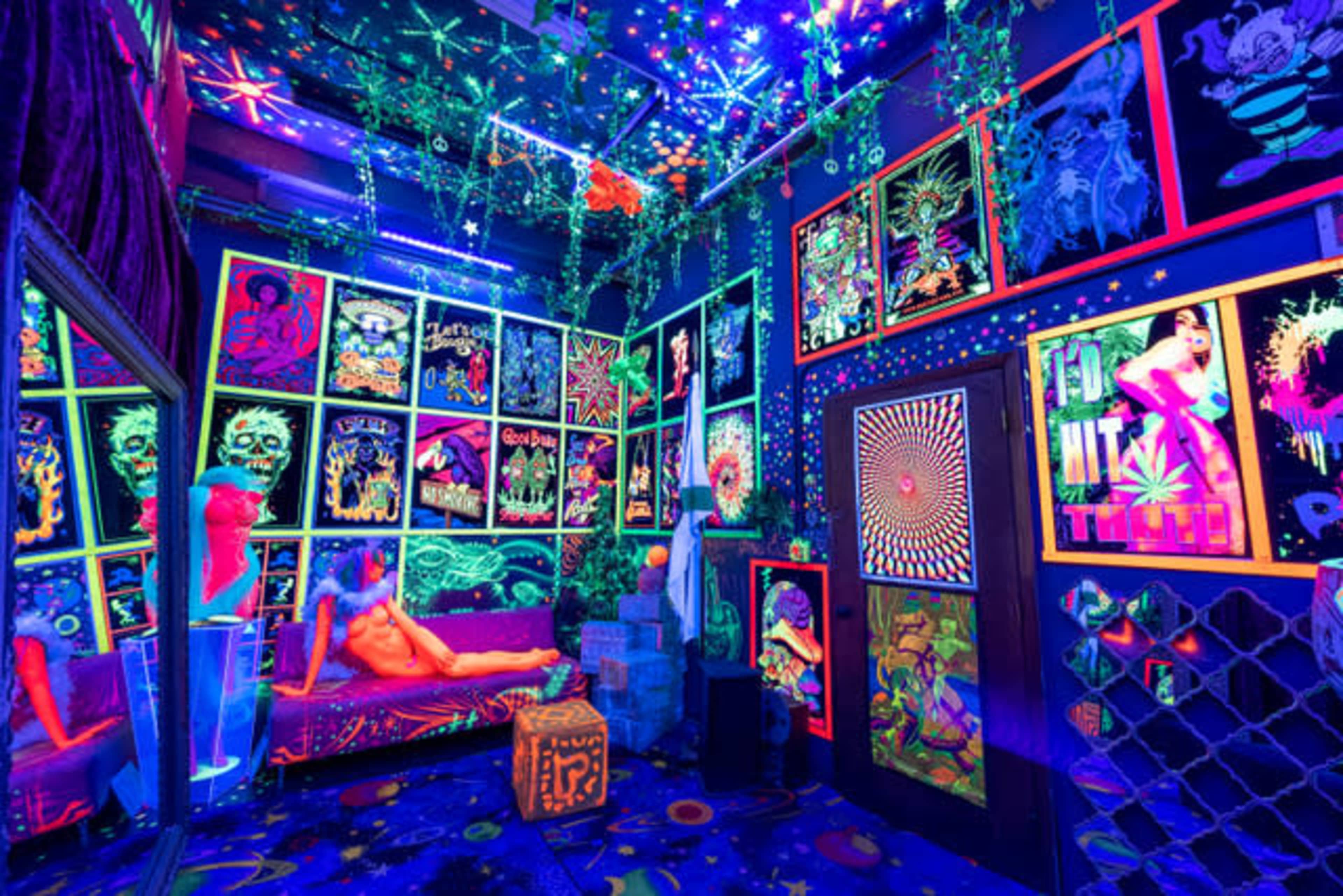 A room decorated with vibrant posters and neon lights, featuring a couch and various art pieces under blacklight.