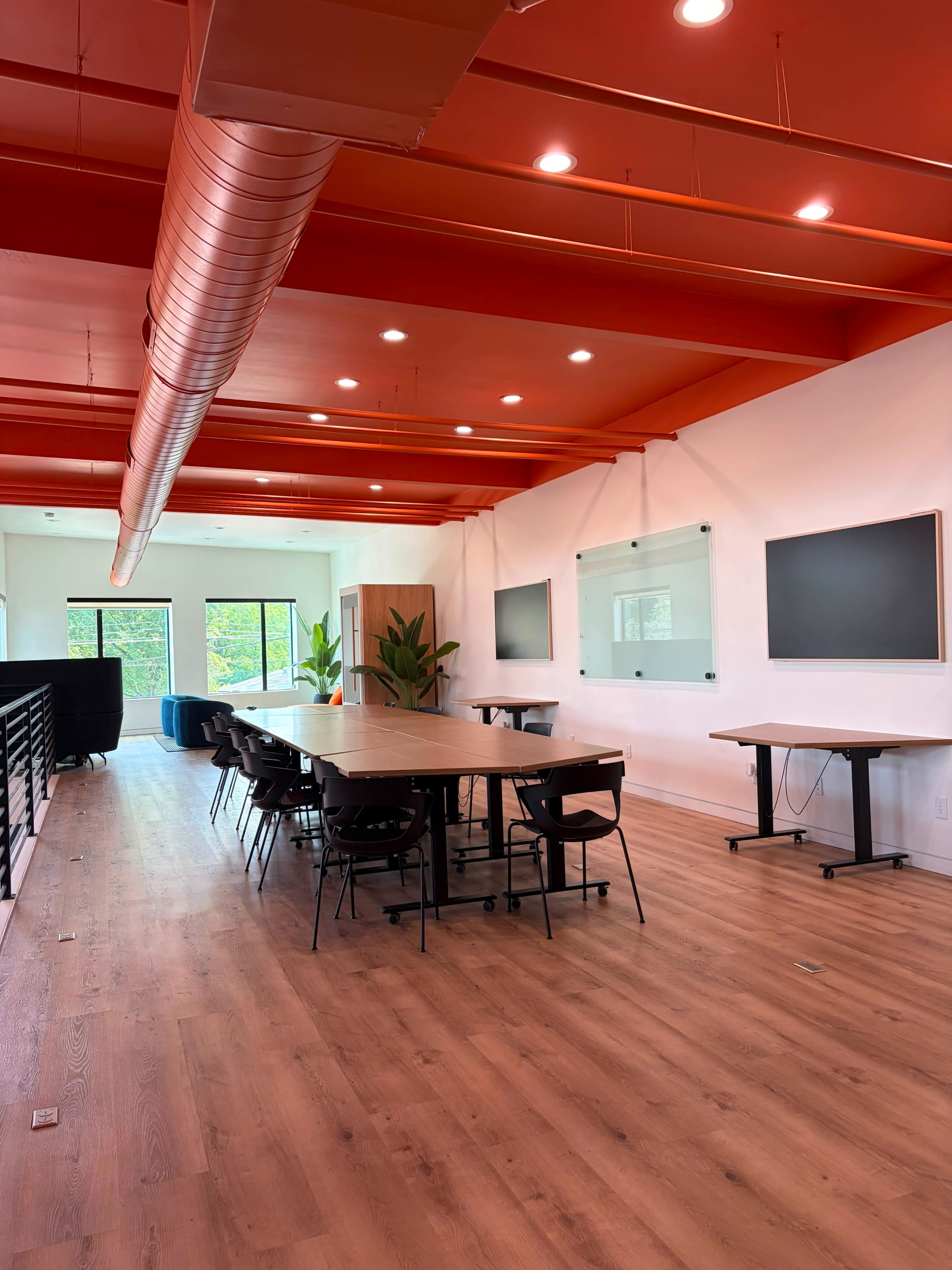 The image shows a spacious conference room with a large wooden table, black chairs, and two wall-mounted screens, all set against a backdrop of red ceiling beams and large windows.