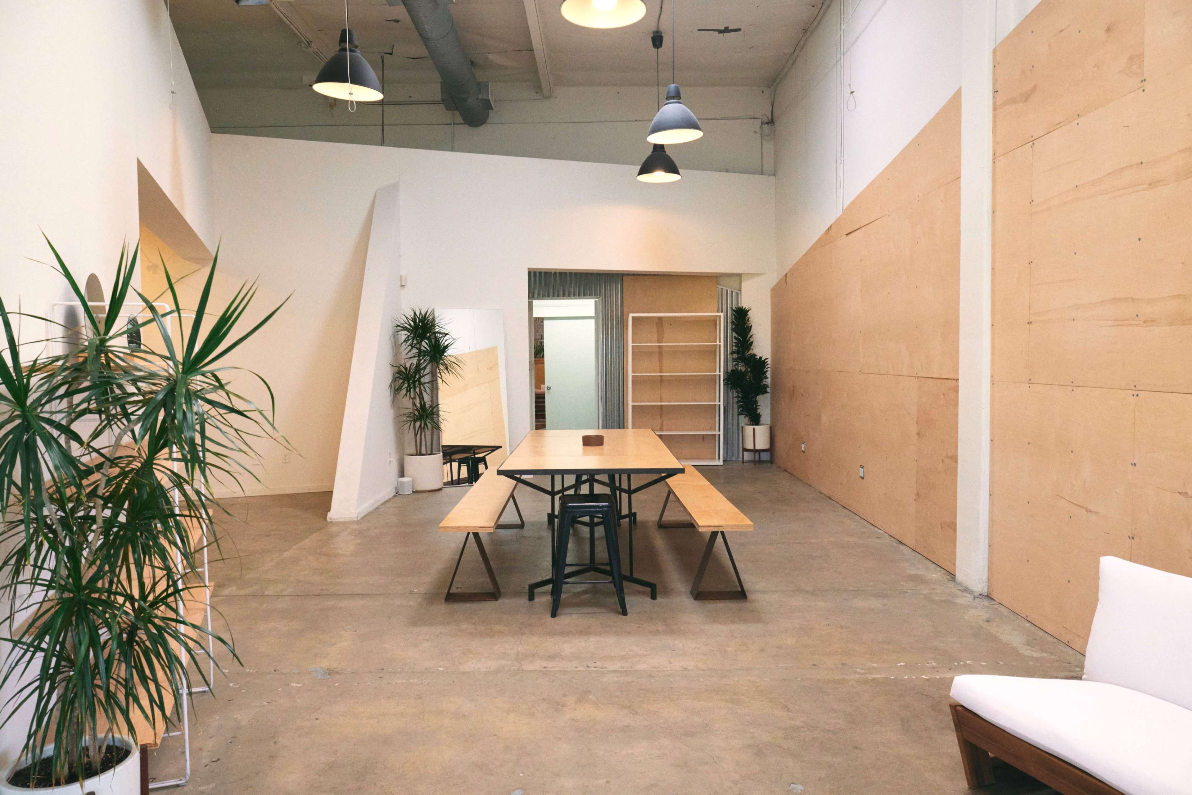 A spacious, minimalist meeting room featuring a large wooden table surrounded by chairs, with potted plants and plywood panels on the walls.
