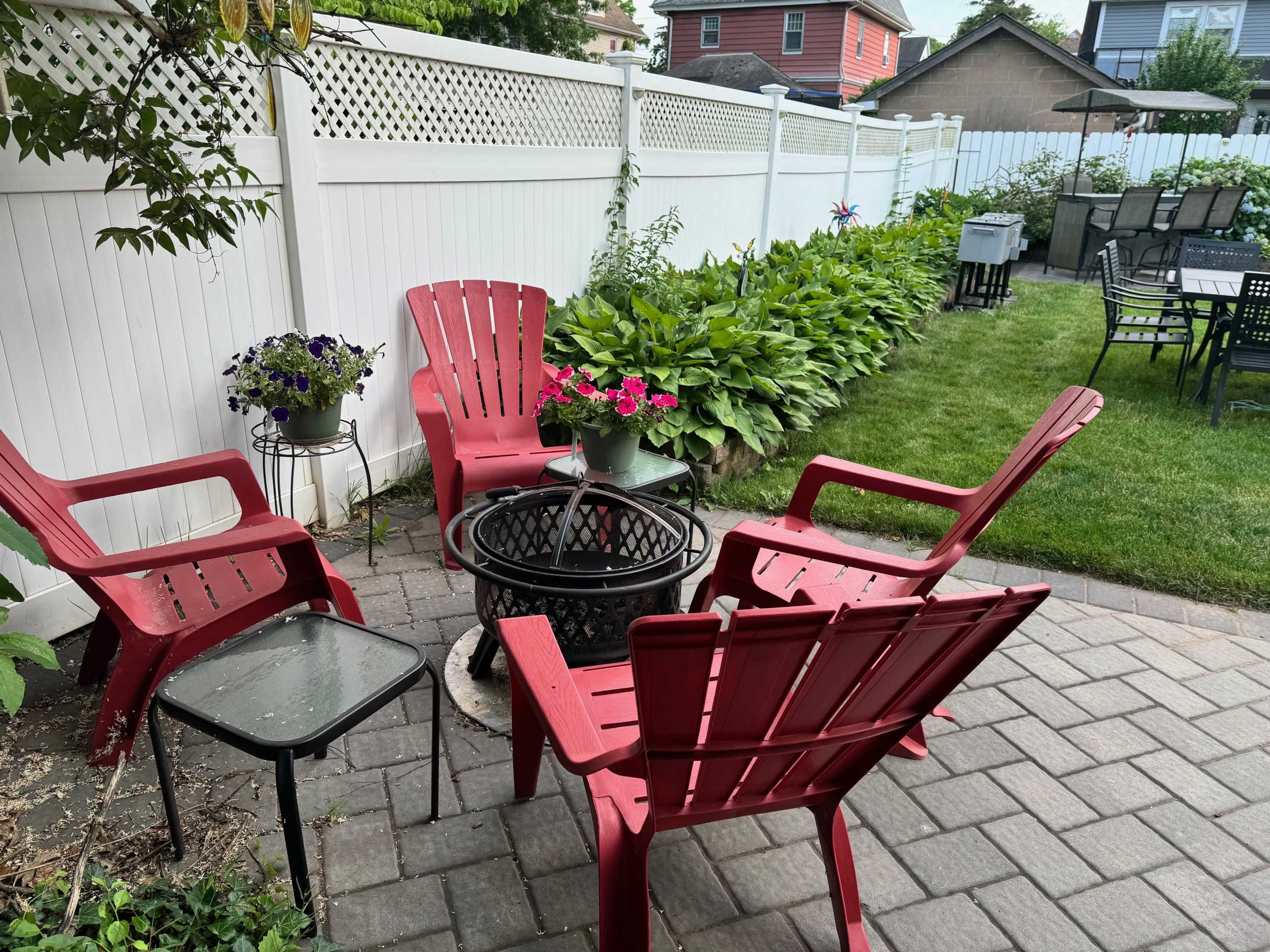 A small patio area features four red Adirondack chairs surrounding a black metal table, bordered by a flower bed and a white fence.