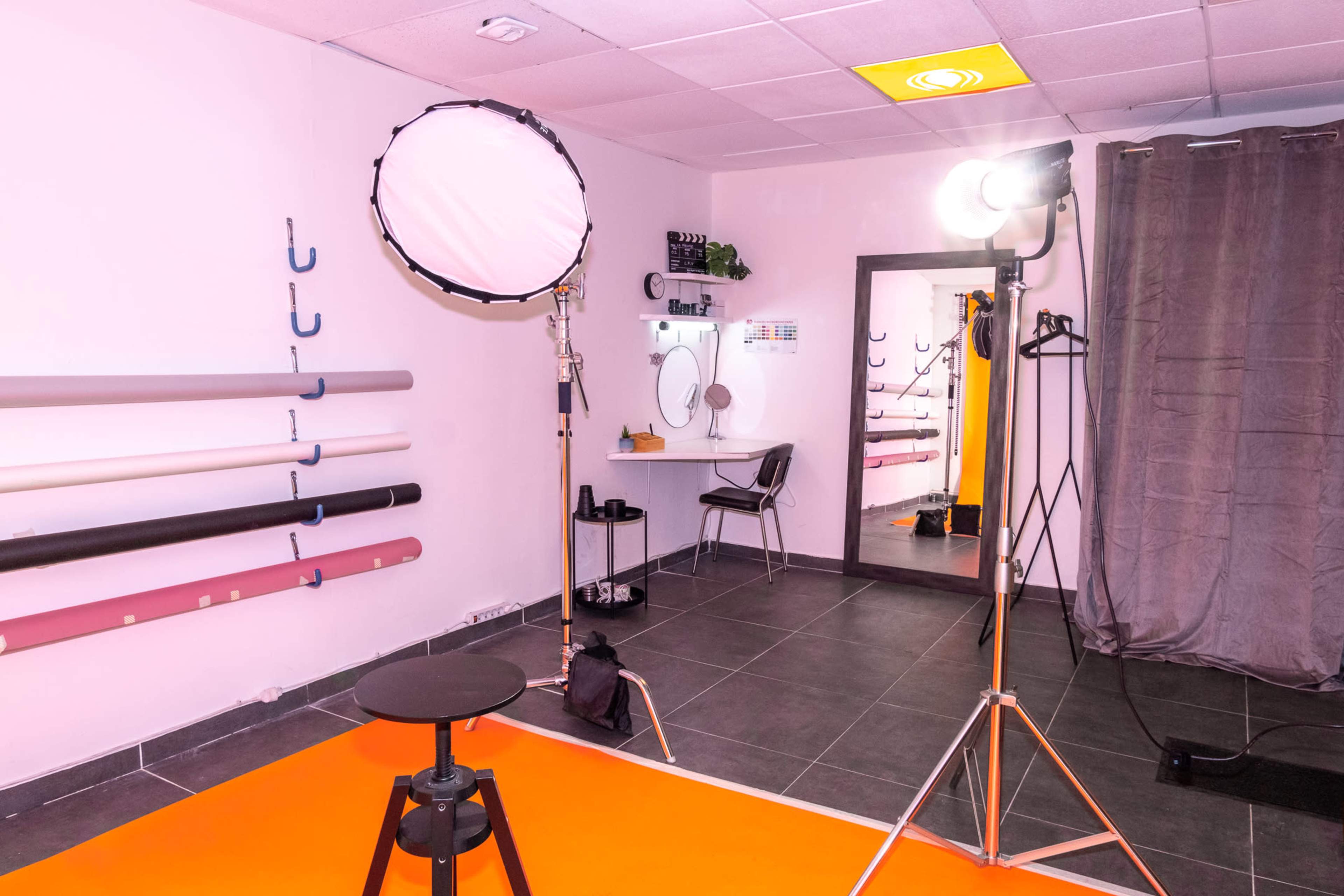 A photography studio with a single chair, lighting equipment, a mirror, and various rolls of backdrop materials displayed on the wall.