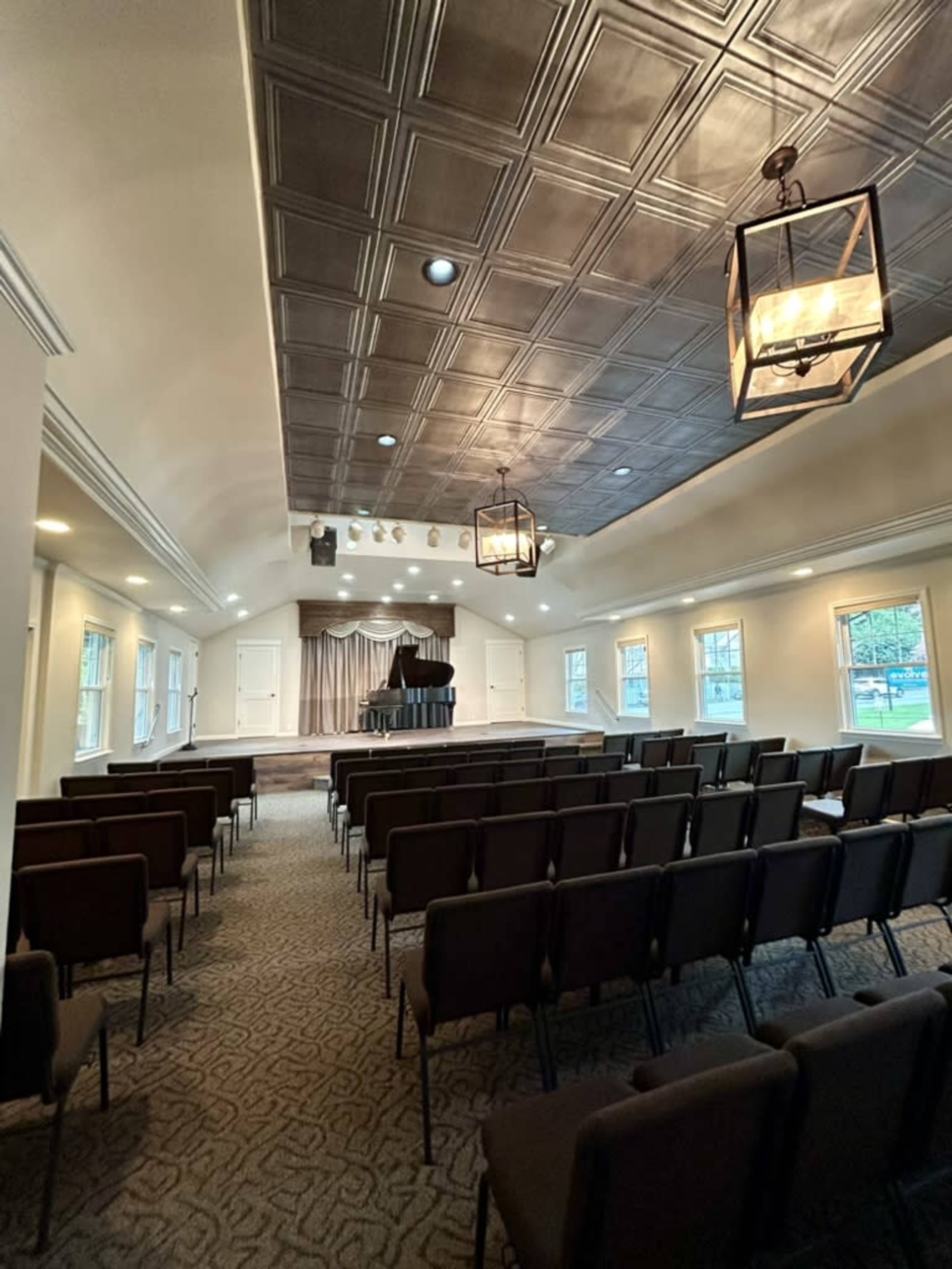 The image shows a neatly arranged auditorium with a grand piano on stage, illuminated by overhead lights and equipped with folding chairs for an audience.