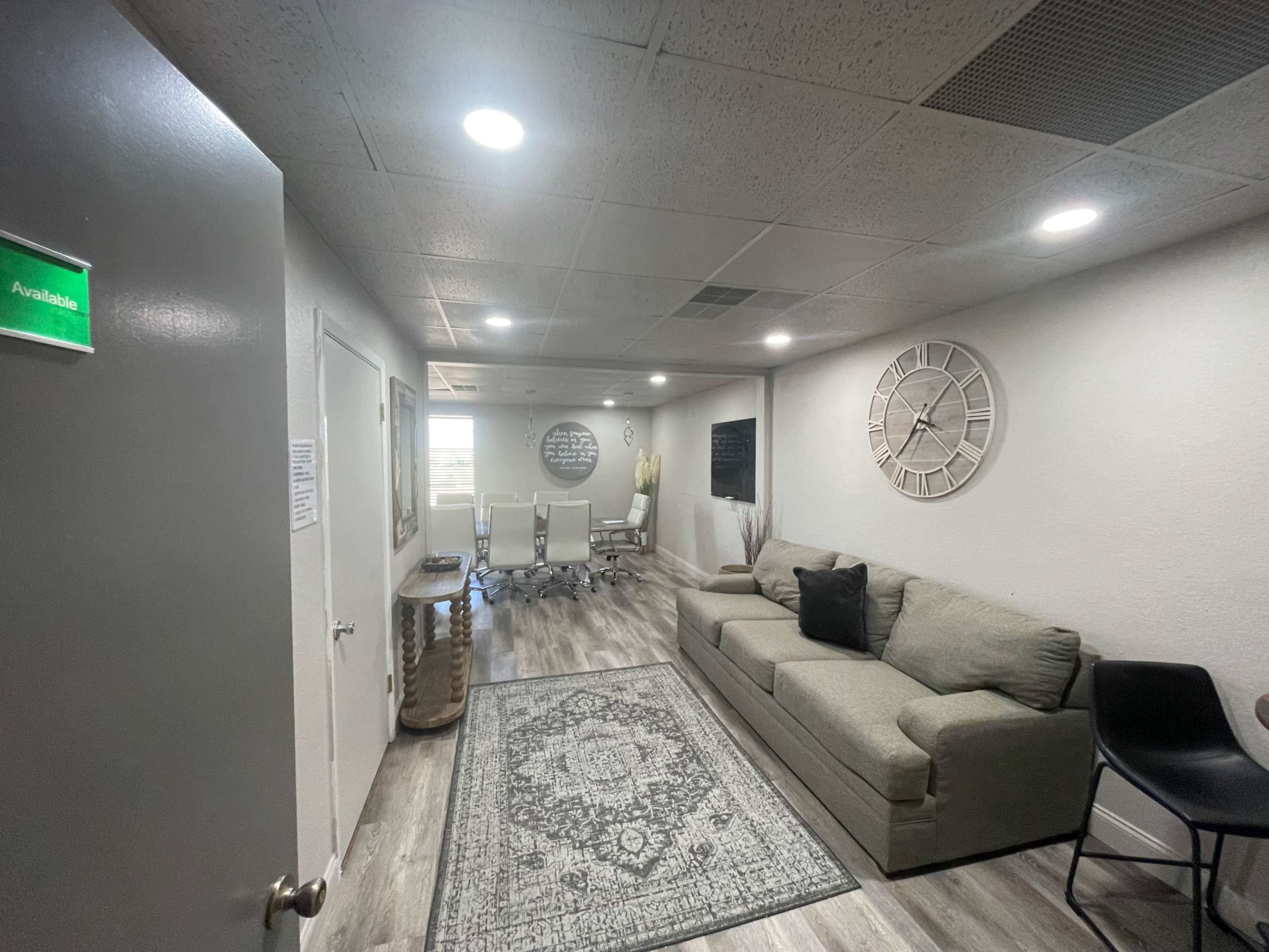 The image shows a modern office space with a gray sofa, a patterned rug, a large wall clock, and a glass conference table in the background.
