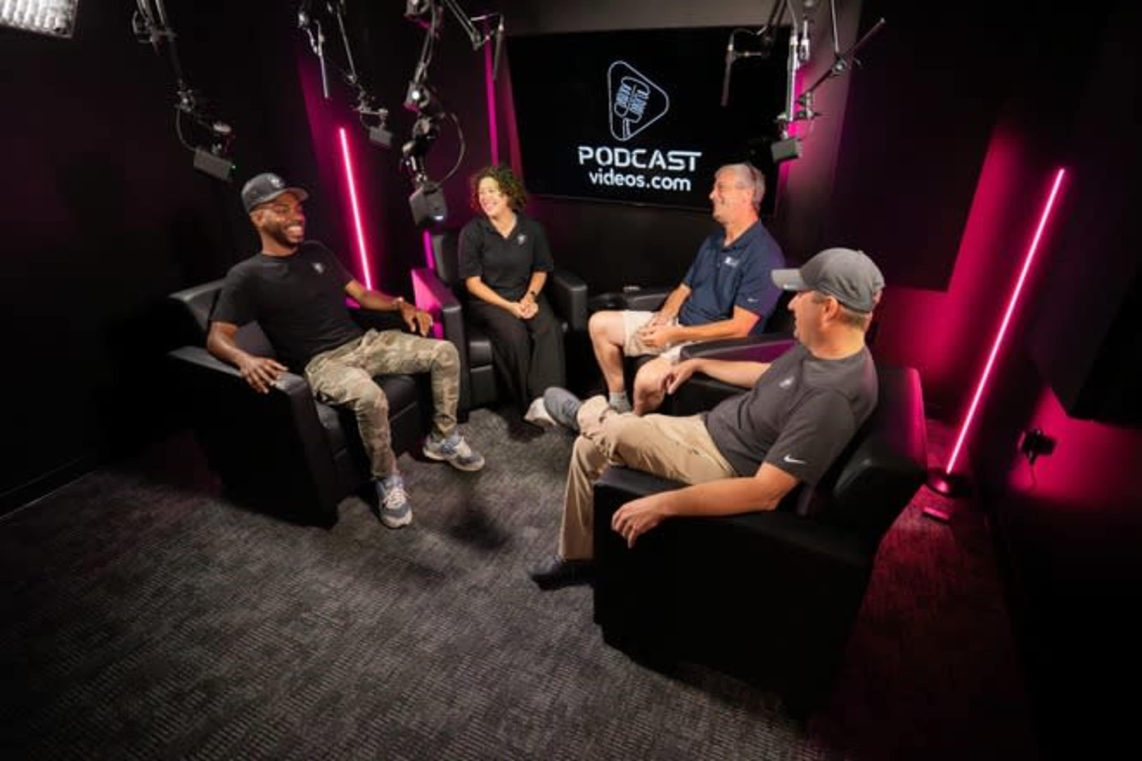 Four people are seated in a well-lit podcast recording studio with dark walls and neon accents, engaged in a discussion.