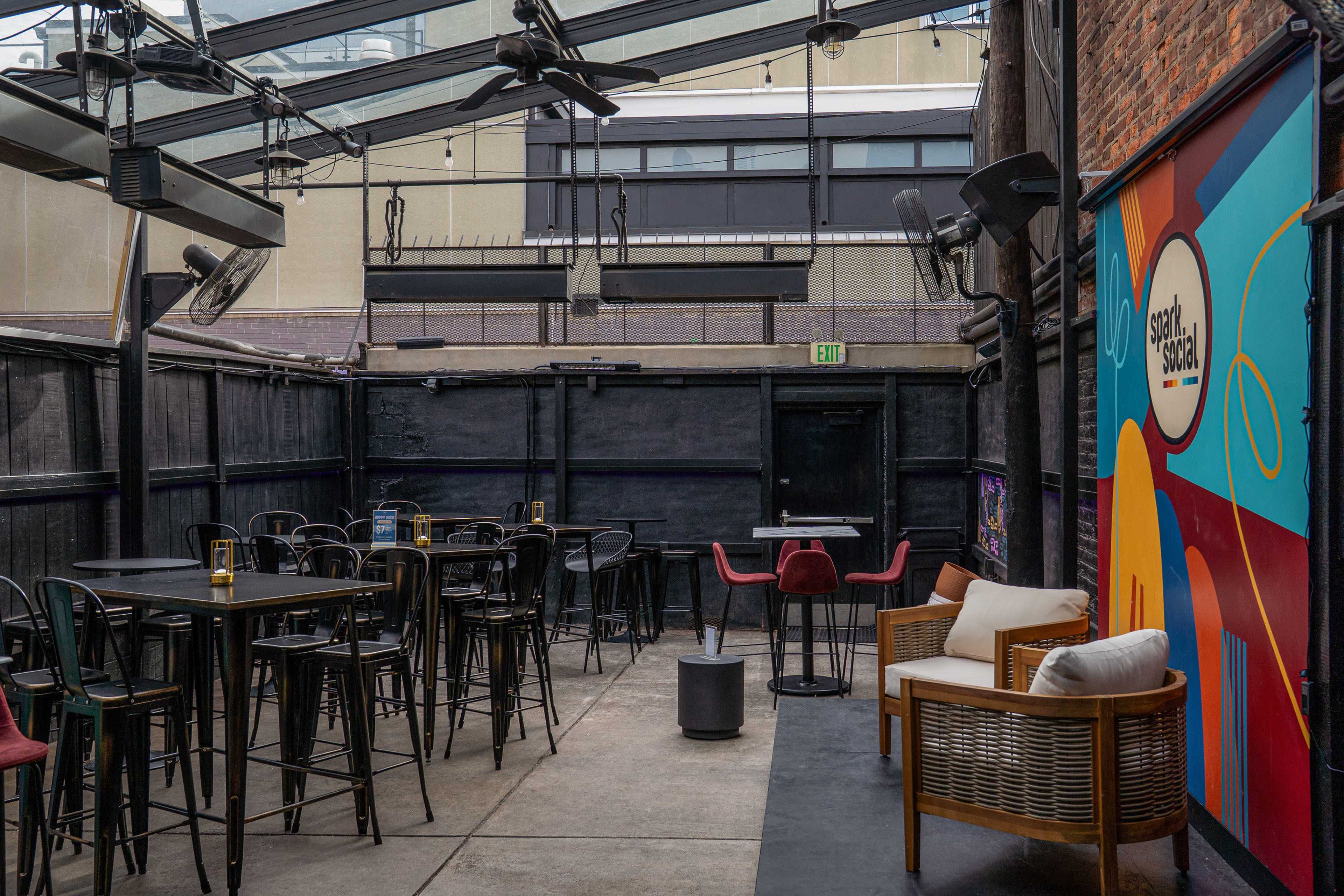 The image shows a spacious outdoor patio area of a bar, featuring high tables and chairs, a few lounge seats, and colorful wall art.