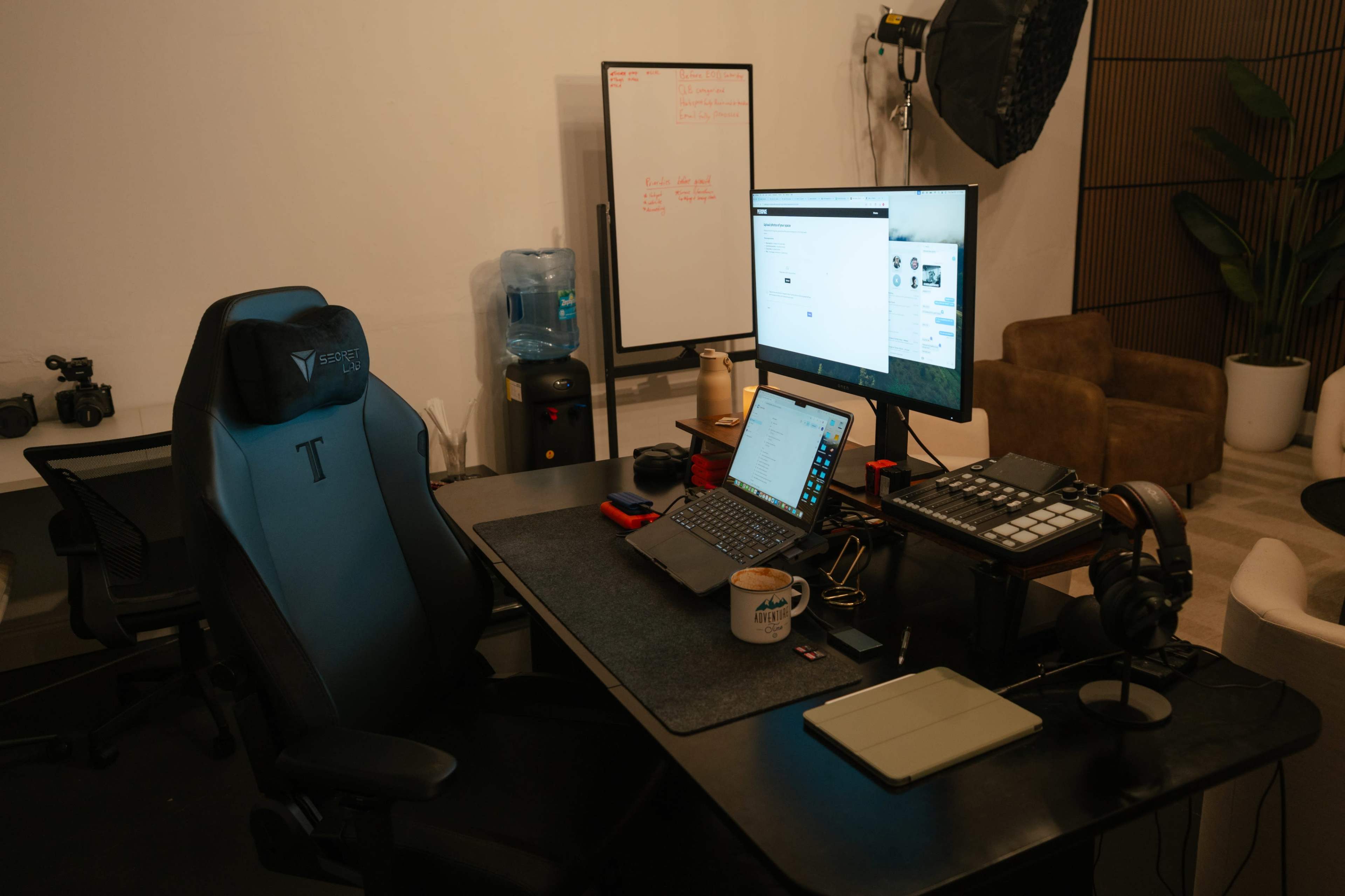 The image shows an organized workspace featuring a desk with two computer monitors, a laptop, a headset, and a water cooler in the background.