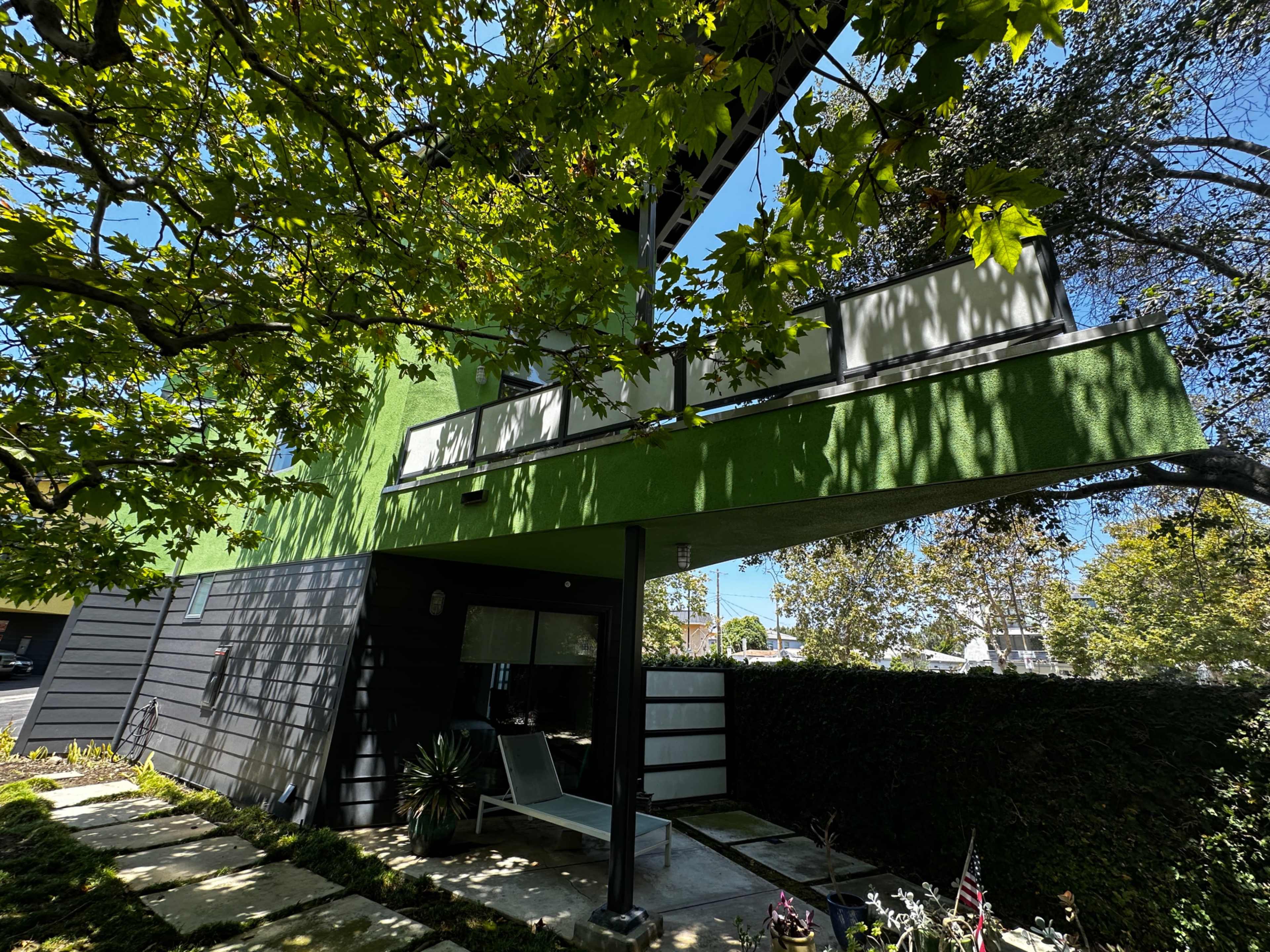 A modern two-story house with a green accent wall is partially shaded by trees and features a patio in the front.